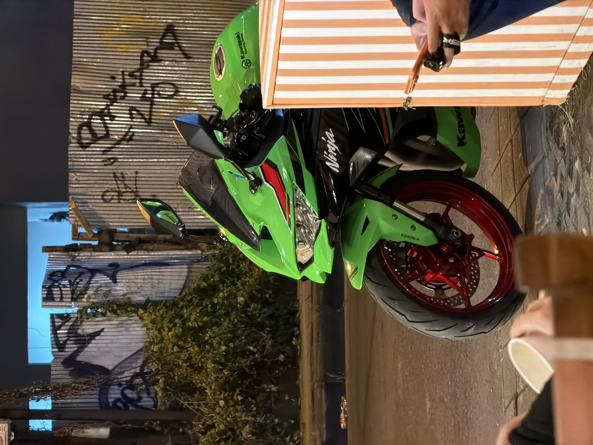The image shows a green Kawasaki Ninja motorcycle parked on a street with light-colored pavement in what appears to be North Kuta, Indonesia. The motorcycle's front tire is red, and the word "Ninja" is visible on the side. To the left, there is a corrugated metal wall covered with graffiti and some green foliage. At the top right, a person is holding keys. The person's ring is visible. The scene is taken from an elevated perspective. At the bottom right of the image, a person is holding a cup, possibly a coffee cup.