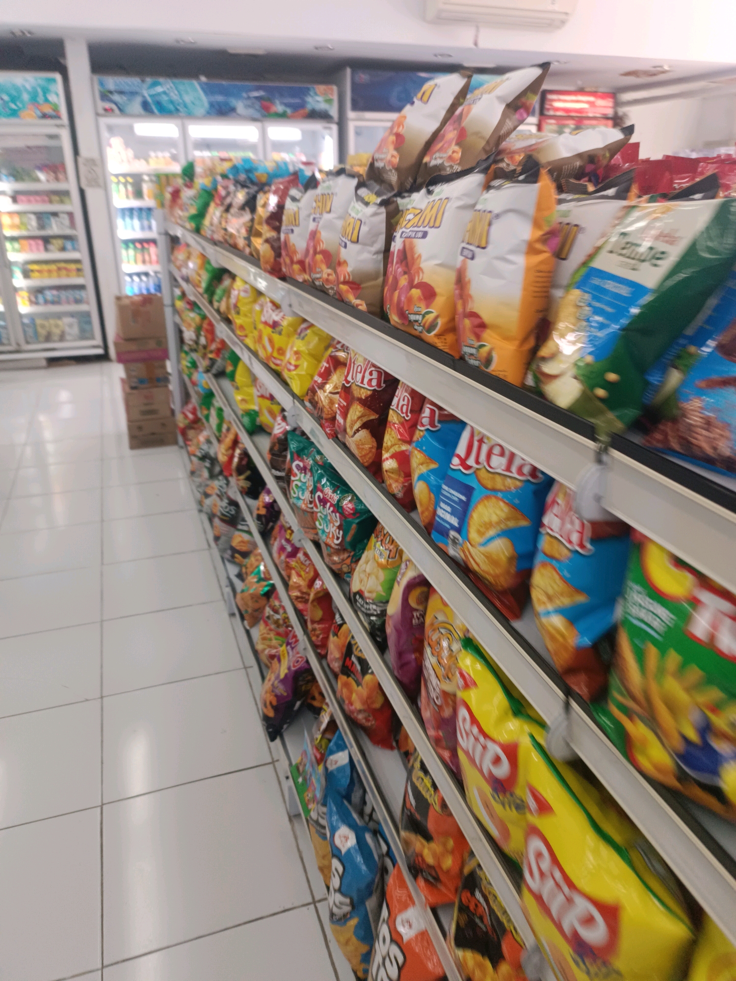 The image shows an aisle in a convenience store in Tabanan, Indonesia, filled with shelves of snacks and beverages. The shelves are stocked with a variety of bagged snacks, including chips and other fried snacks, in colorful packaging. Some of the brands visible include "Letea," "Siip," and "Tempe." In the background, refrigerators stocked with drinks are visible, along with cardboard boxes stacked on the floor. The floor is tiled with white squares. The lighting suggests an indoor environment, likely a brightly lit store. There are no people visible in the image. The perspective is from a low angle, looking down the aisle.