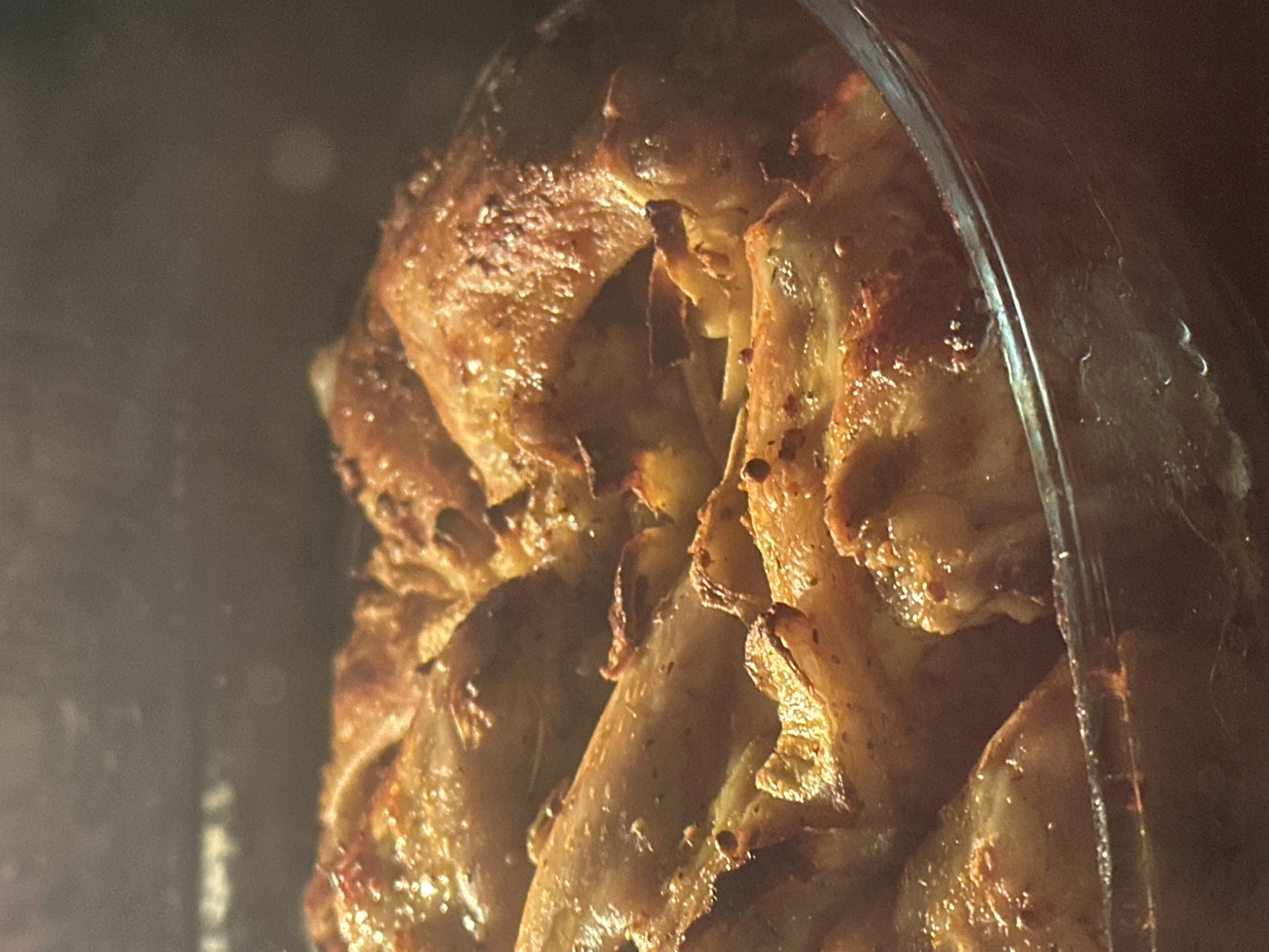 A close-up shot features a large piece of golden-brown, seasoned meat, likely chicken or another poultry/meat cut, being cooked on a rotisserie. The meat has a rich, crispy texture with visible spices and an inviting sheen, suggesting it is succulent and actively roasting. It is positioned vertically, supported by what appears to be a rotisserie spit or central rod, which is partially visible.
The scene is set within an enclosed cooking appliance, indicated by a curved, transparent glass or plastic barrier on the right side of the frame. This barrier shows reflections and possibly some condensation or drippings from the cooking process. The background is dark, blurry, and out of focus, drawing the viewer's attention entirely to the cooking meat.
The warm, artificial lighting enhances the appetizing colors and textures of the food. No people are visible, nor is any discernible text. The image captures the delectable process of meat preparation, evoking a sense of savory cuisine, potentially from the Budva, Montenegro region where such grilled or roasted dishes are popular.