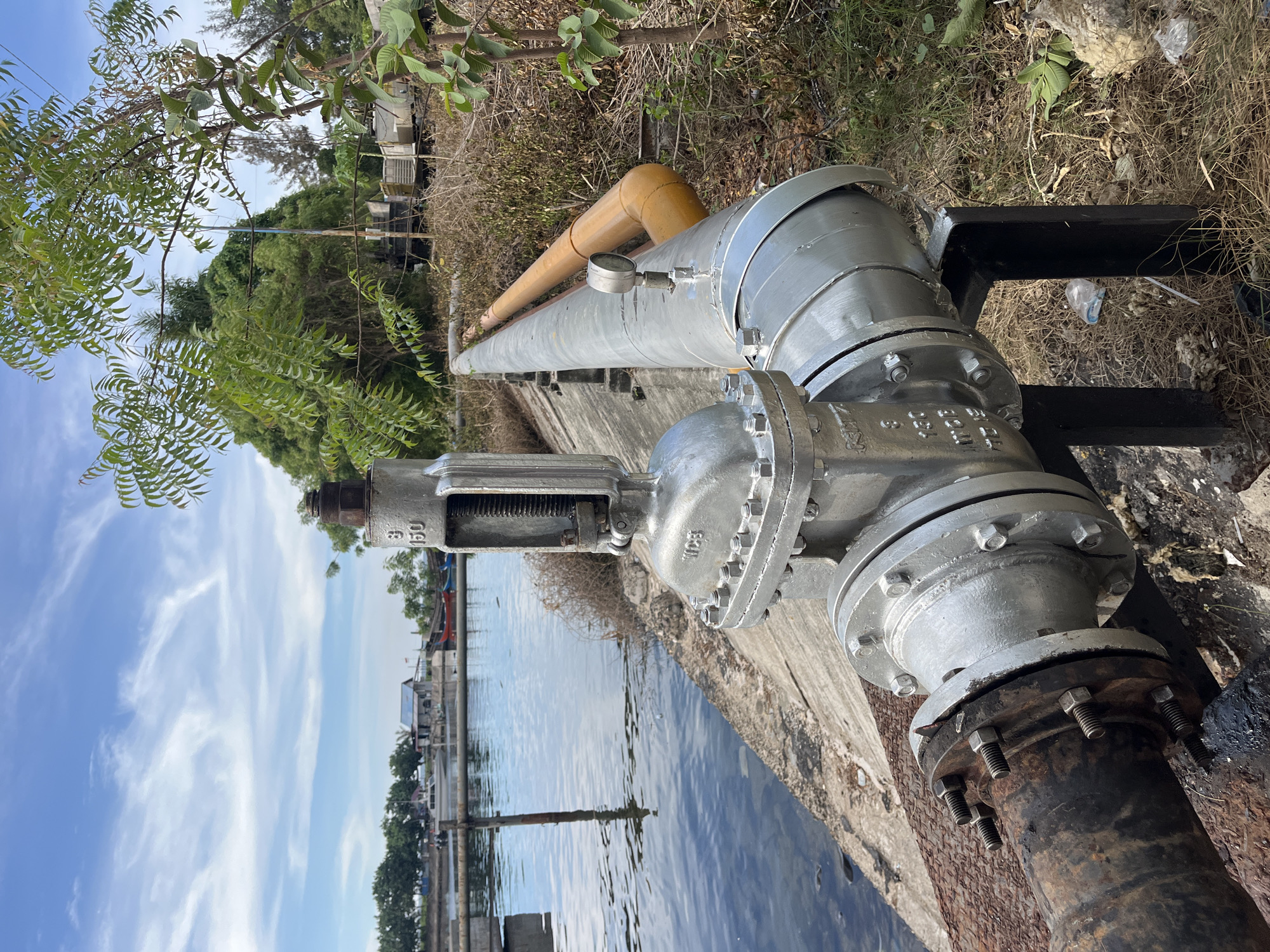 The image shows a large, silver-painted industrial valve and pipeline assembly situated on a concrete ledge adjacent to a body of water. The valve is connected to a dark, rusted pipe extending down towards the water. Another pipe runs horizontally along the ledge, with a yellow pipe positioned above it. Vegetation grows alongside the pipes, and in the background, structures and trees are visible across the water. The sky is blue with scattered clouds. The location is in Tambonbaroh, Indonesia.