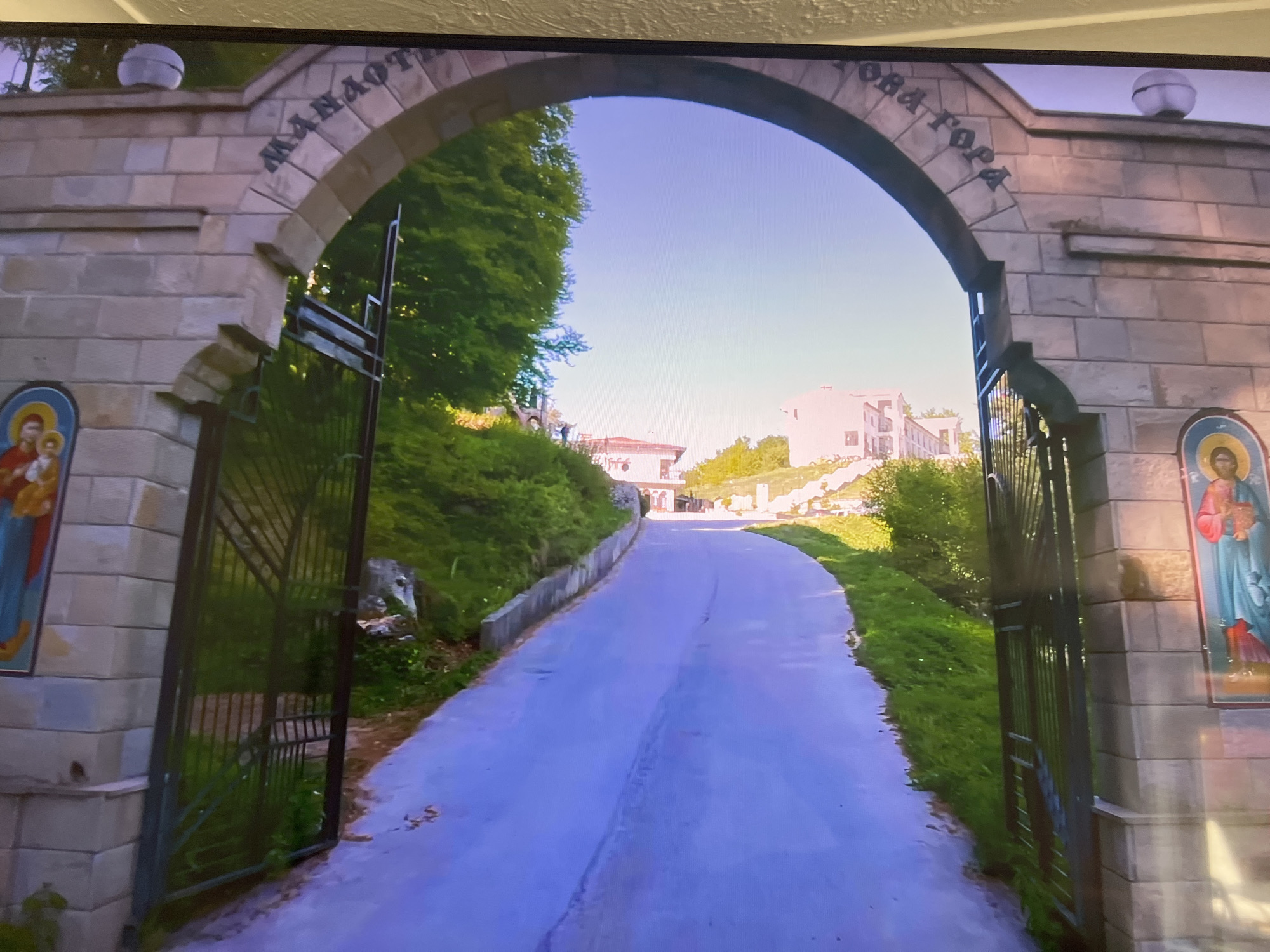 The image shows a view through an arched gateway leading into what appears to be a monastery or religious complex. The gateway is constructed of light-colored stone blocks and has an inscription in Cyrillic script above the arch. The open gates, made of wrought iron, reveal a paved road leading up a gentle slope. Lush green foliage and trees flank the road on both sides. In the distance, buildings are visible. On either side of the gateway are framed religious icons. The location is Unknown, Unknown.