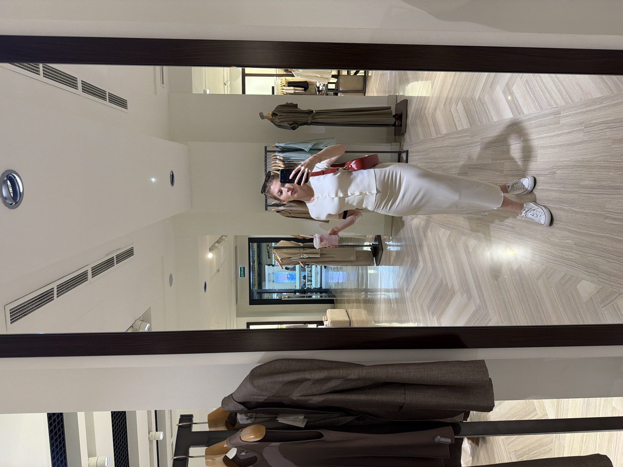 A woman is taking a picture of herself in a mirror inside a clothing store. She is wearing a white dress and white sneakers, and she has a red purse slung over her shoulder. She is holding a cell phone in her right hand and a pink drink in her left hand. The mirror is surrounded by a dark brown frame. The store has a light-colored floor with a herringbone pattern. In the background, there are clothing racks with various garments. The store appears to be located in Moscow, Russia, based on the "Pivot" sign written in Russian. There is a coat hanging on a rack.