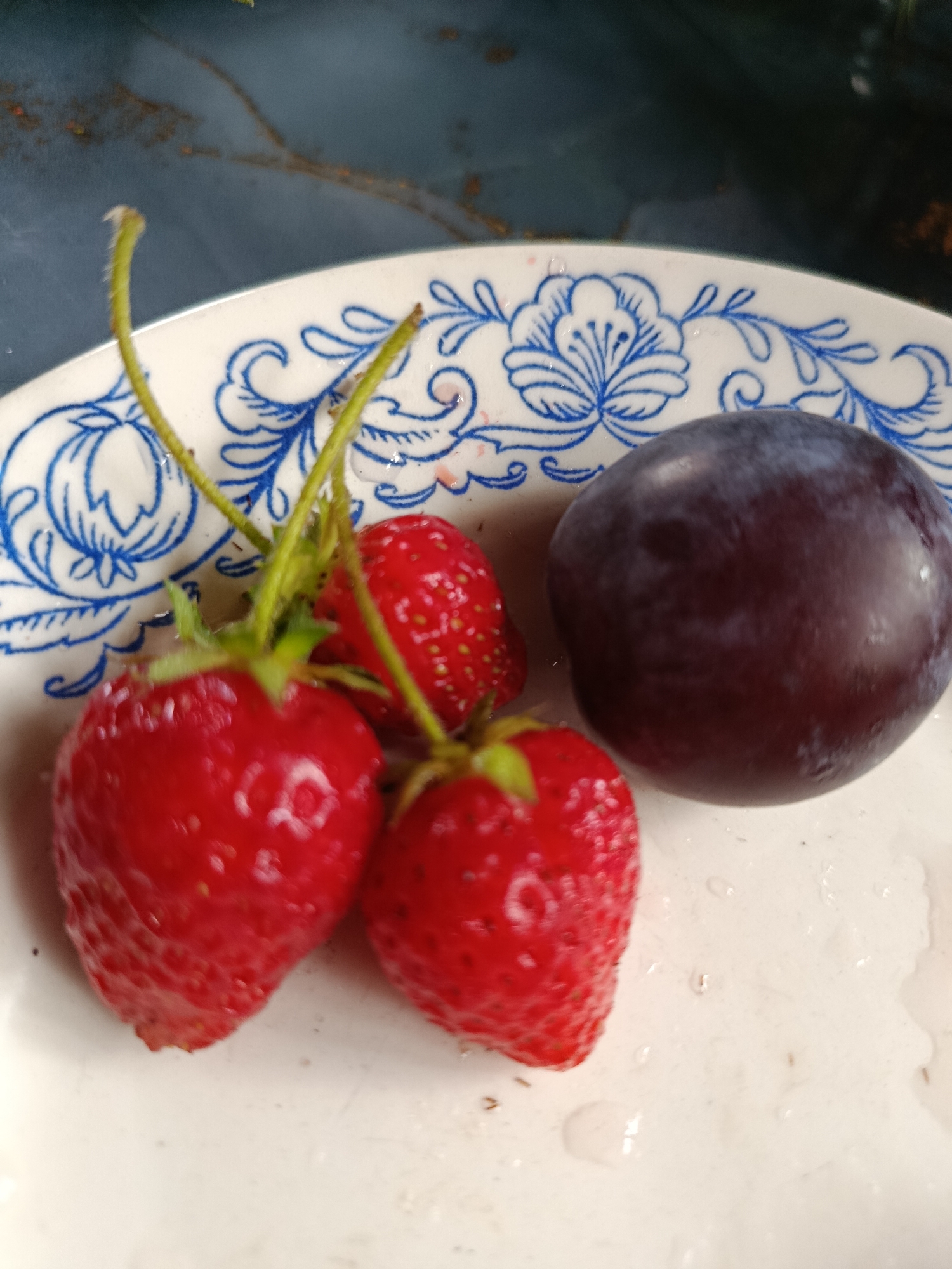 A close-up, overhead shot shows three ripe strawberries and one plum on a white plate with a blue floral pattern. The strawberries are bright red and plump, with their green stems attached. The plum is dark purple and oval-shaped. The plate sits on a dark, textured surface. The image is taken in Verea, Russia.