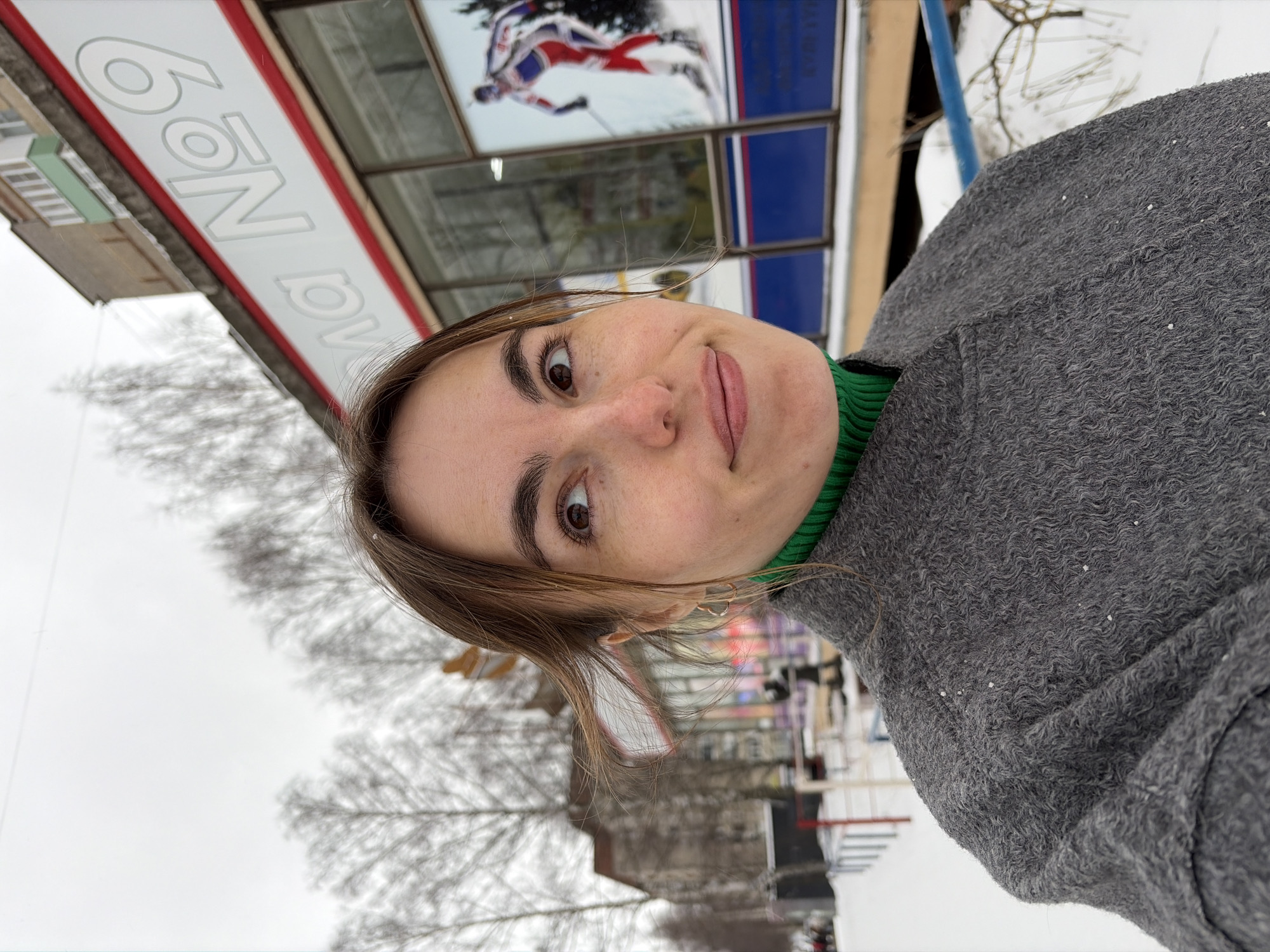 The photograph is a selfie of a fair-skinned woman with brown hair and eyes, wearing a gray coat over a green turtleneck sweater. She is positioned outdoors with a building behind her. The building has a sign that reads "6ON DV" and features a poster of a skier in action. Based on the sign "6ON DV", the location is likely in Izhevsk, Russia, as it seems to refer to "6 on Dvorzovaia" (translated roughly as "6th on Dvorzovaia Street") and Dvorzovaia Street is located in Izhevsk. The environment appears to be snowy, with bare trees and buildings visible in the background, suggesting it is winter.