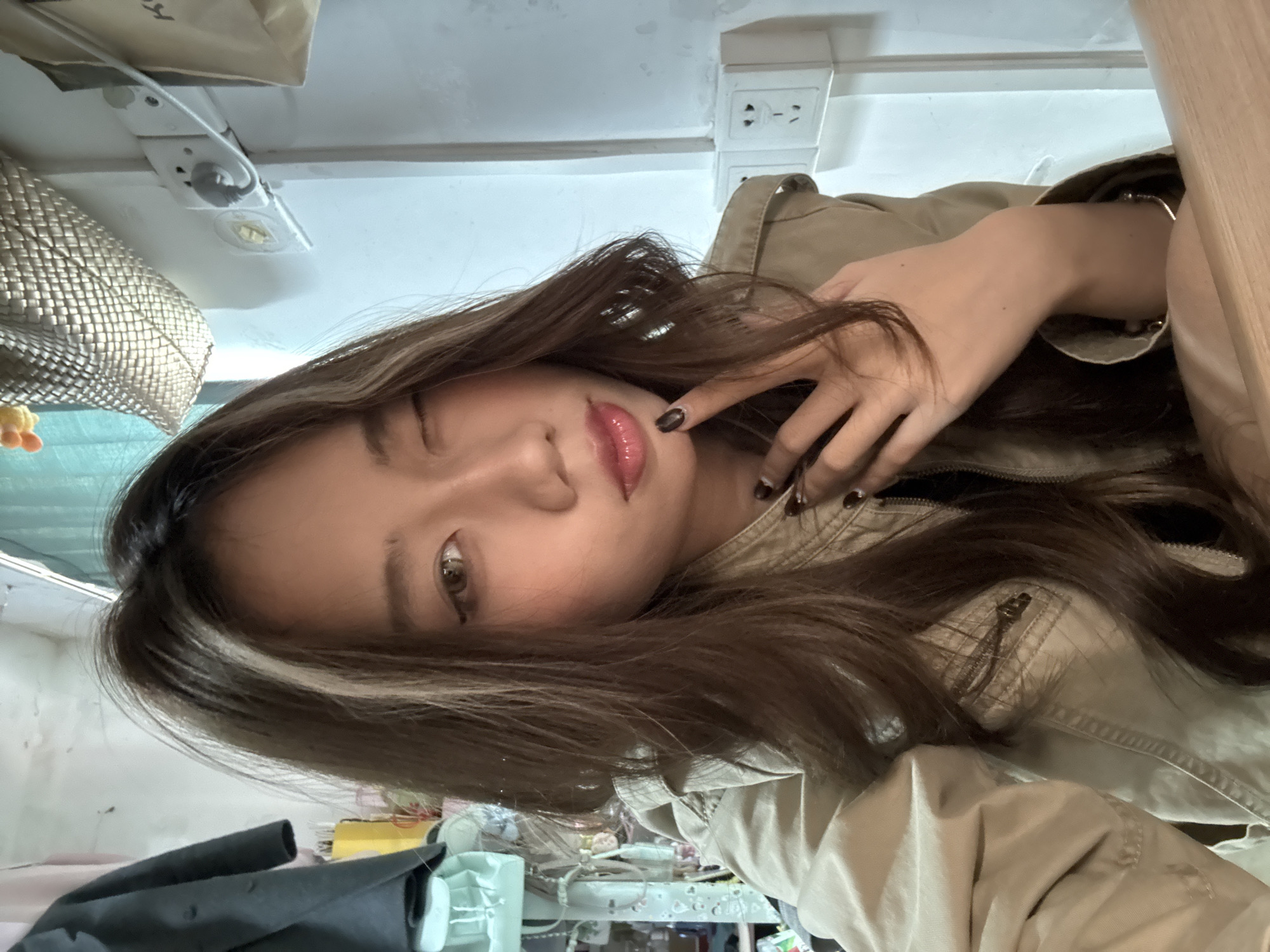 A young woman poses for a selfie in an indoor environment. She has brown hair with a blonde streak, light skin, and is wearing a light brown jacket. Her fingers are near her face, as if posing. In the background, there is an electrical outlet, a light fixture, a garment bag, and items on a table including a black garment. It is not possible to confirm the location in Nan'an District, China from the image.