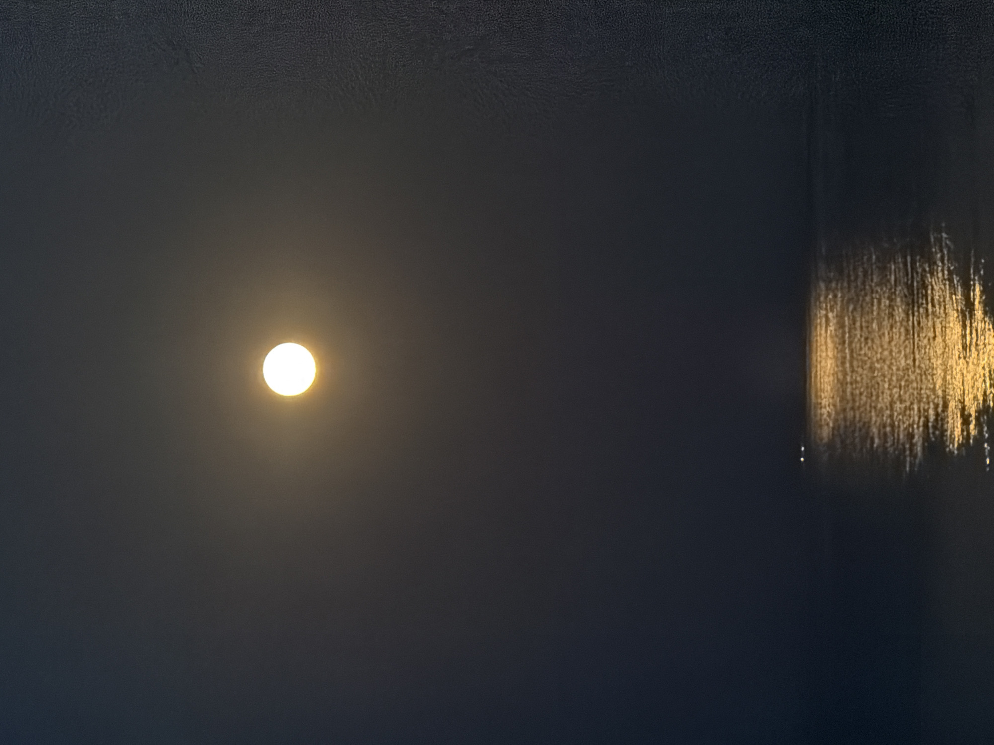 The image depicts a nighttime scene over a body of water, likely the sea, near Νέα Καλλικράτεια, Greece. A bright, full moon hangs in the dark sky, casting a shimmering golden reflection onto the water's surface. The reflection extends from the horizon upwards, creating a luminous path. In the distance, two small, indistinct lights are visible on the water, possibly from a boat or a distant structure. The overall atmosphere is serene and tranquil.
