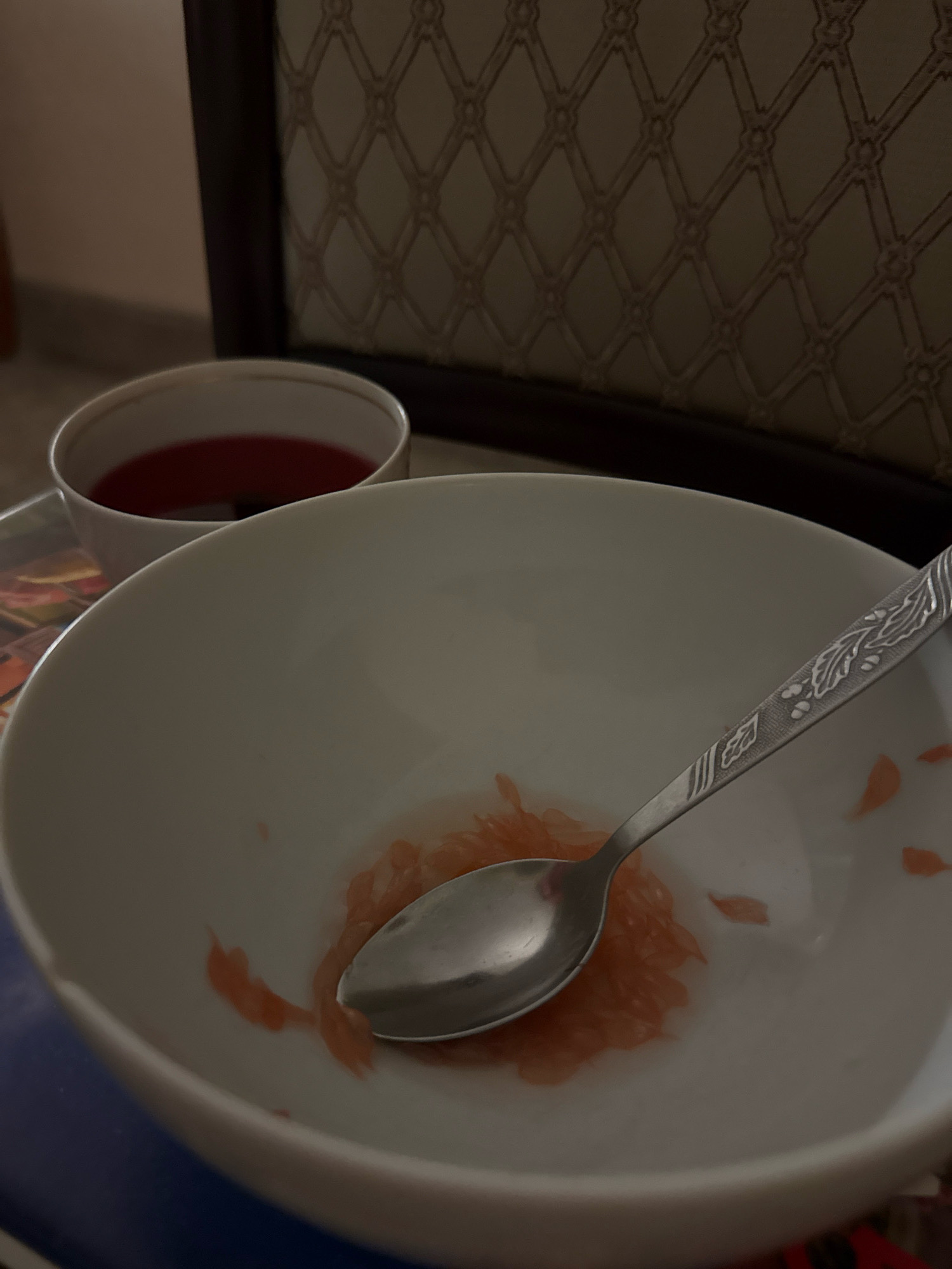 In the city of Zavolzhye, Russia, a close-up shot reveals a white bowl containing the remnants of a meal. A silver spoon rests inside the bowl, with some pink grapefruit pulp adhering to its surface and scattered around the bottom of the bowl. To the left of the bowl, a smaller white cup holds a dark red liquid, possibly juice or tea. The background is blurred, showing a textured surface with a diamond pattern and the edge of a dark wooden frame. The lighting is dim, suggesting an indoor setting.