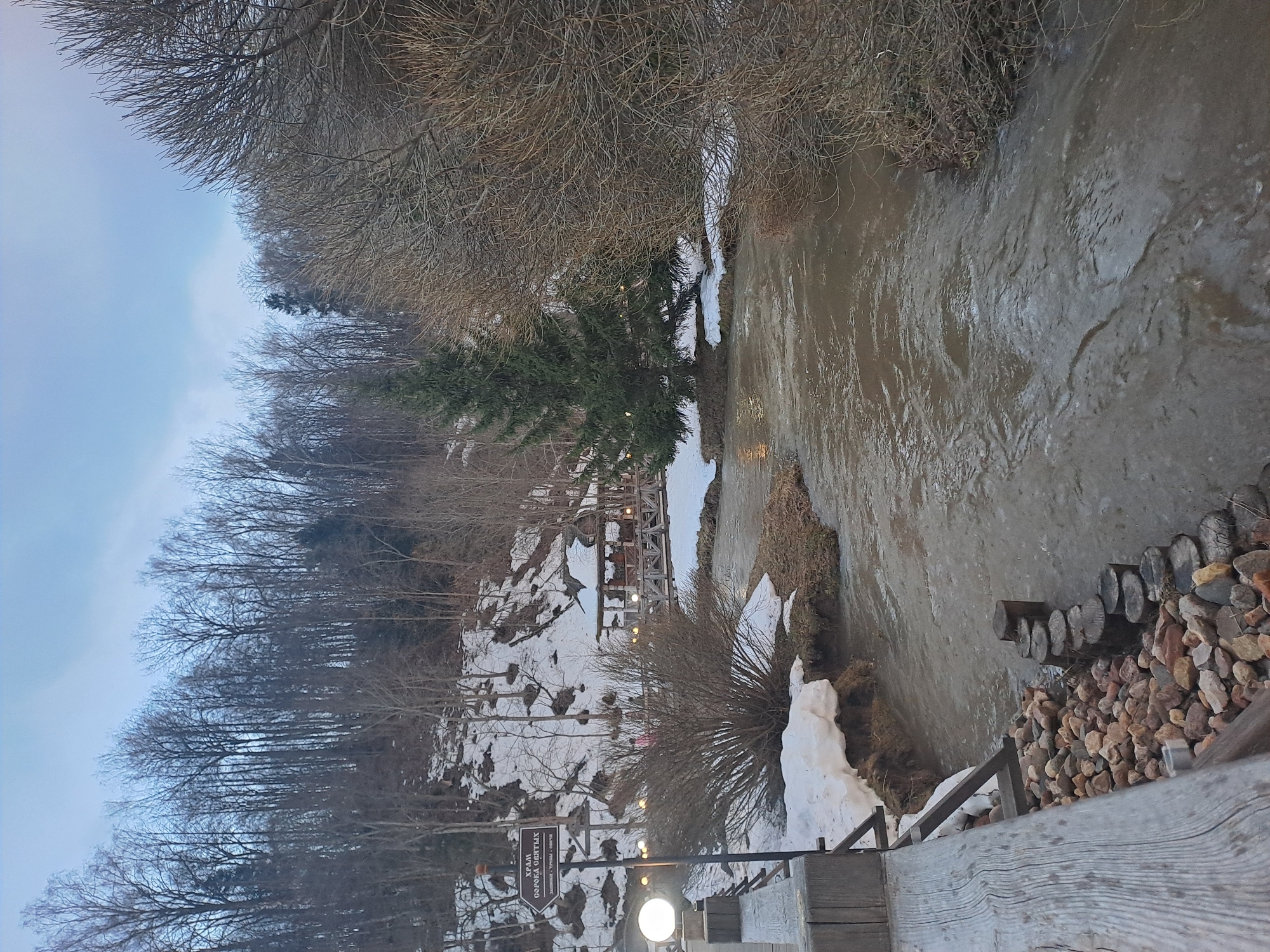 The image depicts a landscape in what appears to be a partially snow-covered area. A body of water, possibly a river or stream with murky water, flows through the scene. The riverbank is lined with vegetation, some of which is bare due to the season. Patches of snow are visible along the banks and on the surrounding landscape. Bare trees and branches dominate the left side of the frame. In the distance, what appears to be a building or structure, possibly a restaurant, is visible on a slope. A wooden railing and rocks are present in the lower right corner, suggesting a viewpoint or pathway. The sign present reads, "Храм Добрка Овитх". The location is Vzglyadnevo, Russia, based on the sign's context.