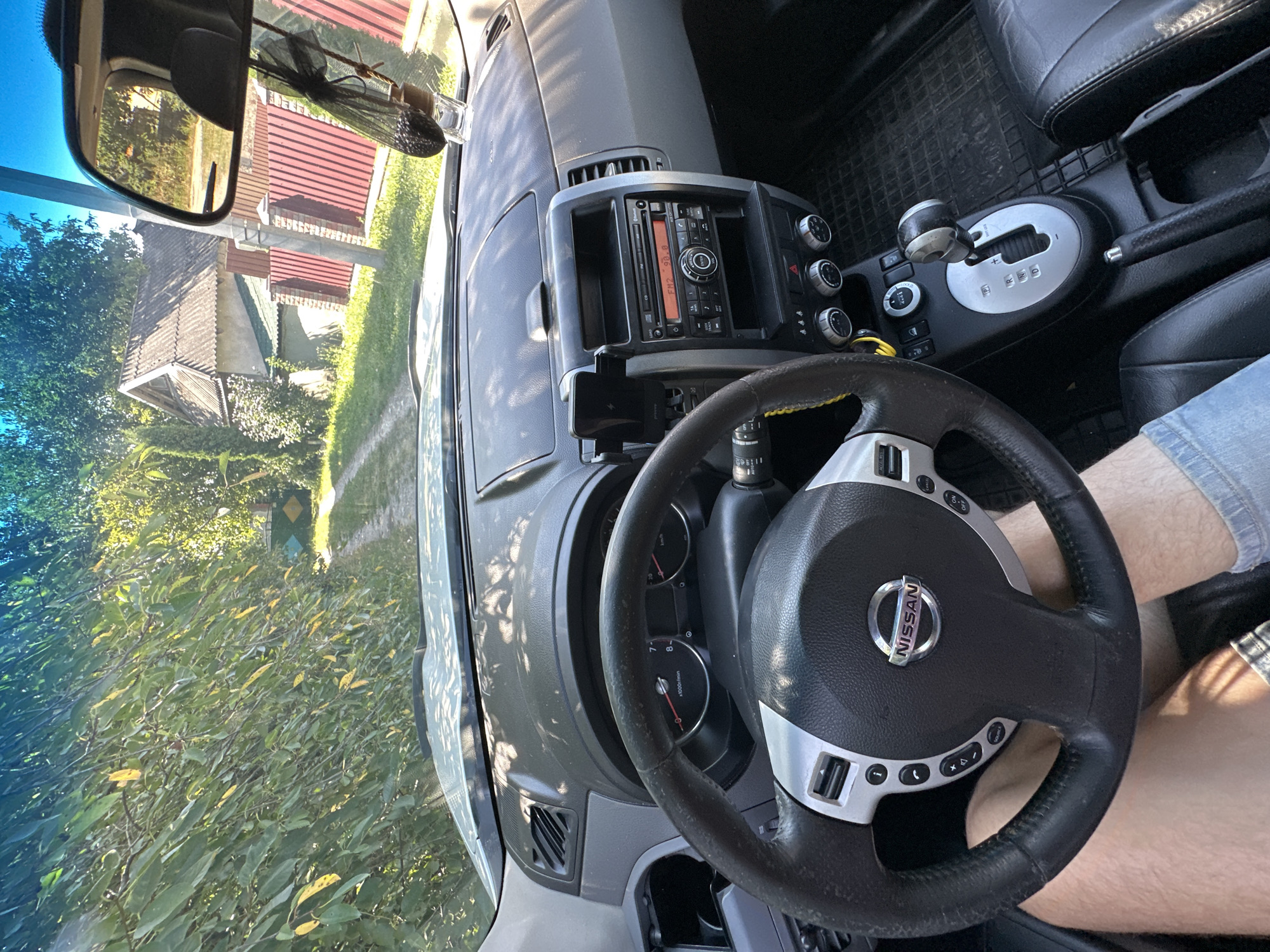 The image is an interior shot of a Nissan car, likely taken in Shatava, Ukraine. The viewpoint is from the driver's seat, looking towards the dashboard and the road ahead through the windshield. The dashboard features a steering wheel with the Nissan logo, a car stereo displaying "FM2 90.0", climate control knobs, and a gear shift. Outside the car, a dirt road lined with trees and bushes is visible, leading towards a rural house with a red-tiled roof and a red fence. The sun is shining, casting shadows on the dashboard. A person's legs are visible in the lower part of the frame.