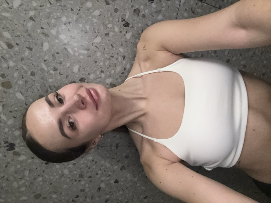 A young adult female is depicted in a close-up, chest-up selfie format, looking directly into the camera with a subtle smile. She has light skin, dark hair pulled back, and features enhanced eyelashes and glossed lips. A small silver hoop earring is visible in her left ear. She is wearing a plain white halter-neck crop top that reveals her midriff. A small, outlined heart tattoo is present on her left shoulder. The subject is positioned against a textured wall surface, uniformly patterned with grey, white, and dark speckled aggregates, resembling terrazzo. The lighting is even. There are no visual cues within the image to determine a specific location in Kyiv, Ukraine.