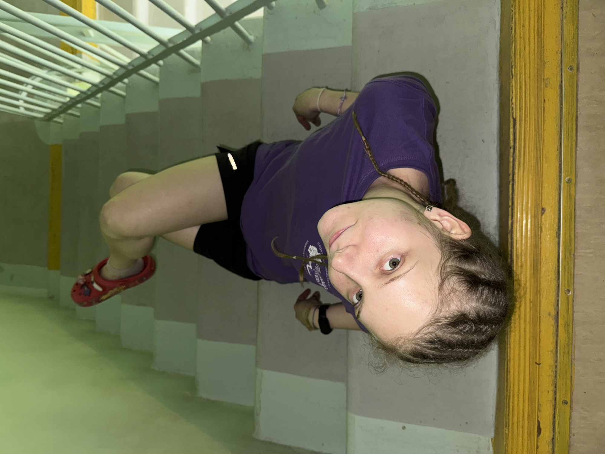 A woman is sitting on a flight of stairs indoors. She is wearing a purple t-shirt, black shorts, and red shoes. The stairwell has a metal railing and the walls are painted in light shades. The location is suggested to be Berezniki, Russia.