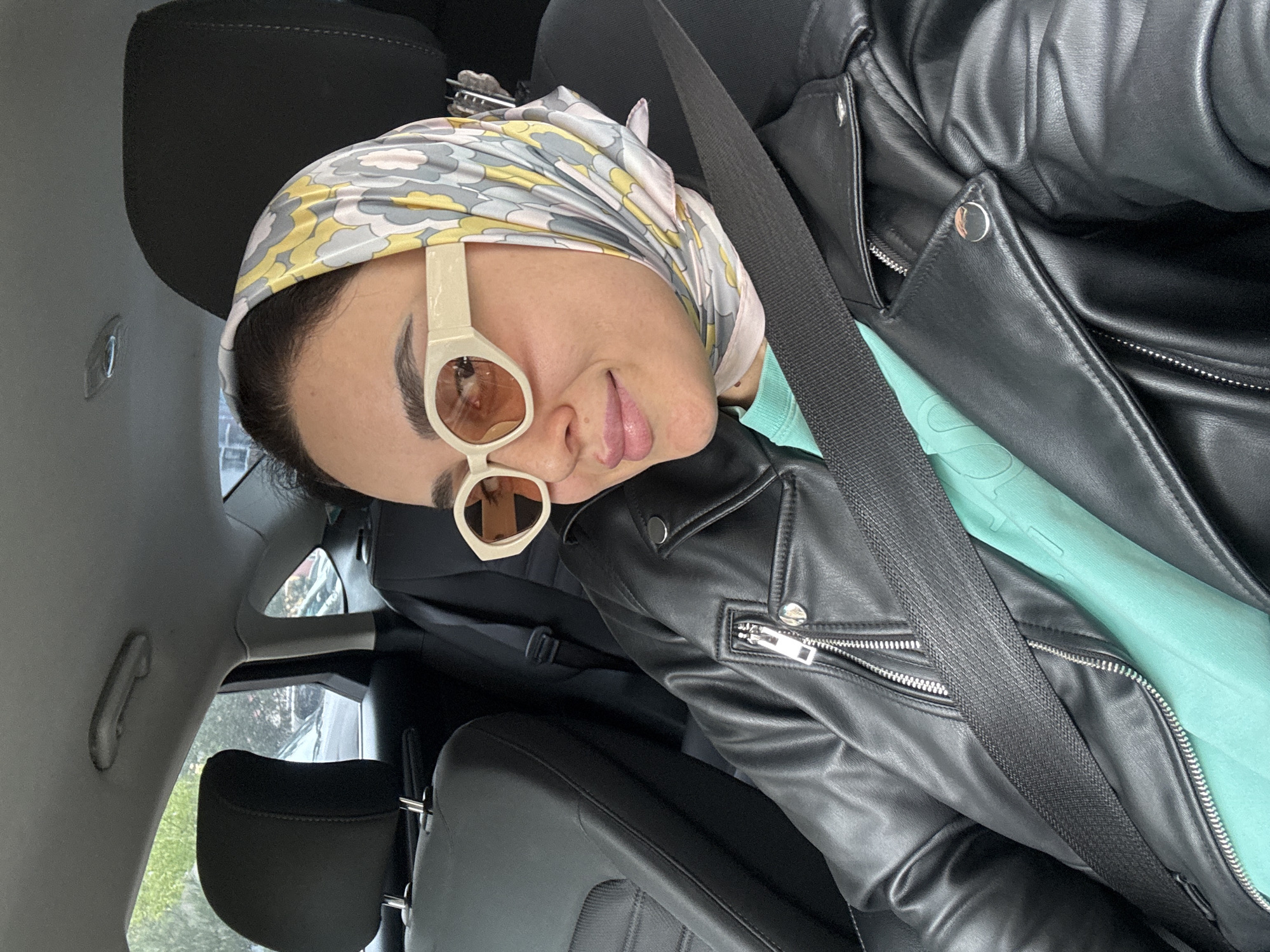 A person is seated in the passenger seat of a vehicle, wearing a black leather jacket, a light blue shirt, a headscarf with a yellow, gray, and white pattern, and sunglasses with white frames and brown lenses. They are wearing a seatbelt. The interior of the vehicle includes black leather seats, a gray interior panel, and a partially visible window showing trees outside. The location is likely in Tbilisi, Georgia.