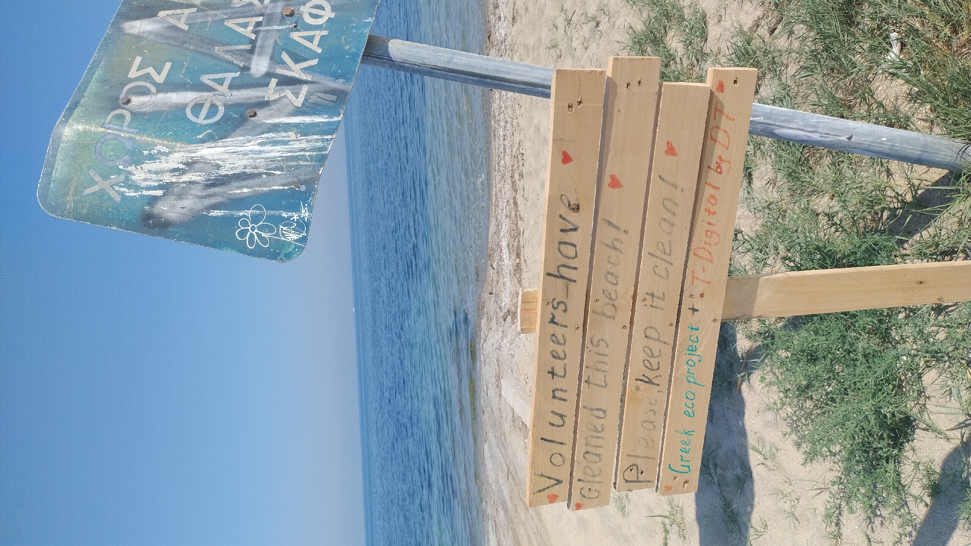 This image captures a beach scene in Thermi, Greece, featuring two distinct signs against a backdrop of a clear blue sky and calm sea.
In the upper right portion of the frame, a weathered, light blue metal sign is affixed to a grey metal pole. The sign is discolored, stained with white streaks resembling bird droppings, and bears white graffiti, including a simple outlined flower. Portions of Greek text are visible, reading "ΧΩΡΟΣ" (Area), "ΘΑΛΑΣΣ" (Sea), and "ΣΚΑΦ" (Vessels), suggesting it indicates a designated area for sea vessels.
Below and slightly to the left of the metal sign, a second sign is constructed from four horizontal light-colored wooden planks, or pallets, attached to a vertical wooden post. Handwritten text in black and colored markers adorns this sign:
* The top plank reads "Volunteers have" in black marker, followed by a small red heart.
* The second plank reads "cleaned this Beach!" in black marker, also with a small red heart.
* The third plank states "Please, keep it clean!" in black marker, accompanied by a small red heart.
* The bottom plank displays "Greek eco project + :" in green marker, followed by "T-Digital by DT" in orange marker, with small red hearts at each end.
The background features a vast, cloudless light blue sky meeting the horizon over the sea. The sea itself is calm, displaying shades of blue and light green near the shore, with minimal ripples. A faint, distant object, possibly a small boat, is visible on the water near the horizon. The immediate shoreline consists of light-colored sand, with some scattered natural debris or seaweed near the water's edge. In the foreground, patches of low-lying green vegetation grow on the sandy ground. The scene is illuminated by natural daylight.
