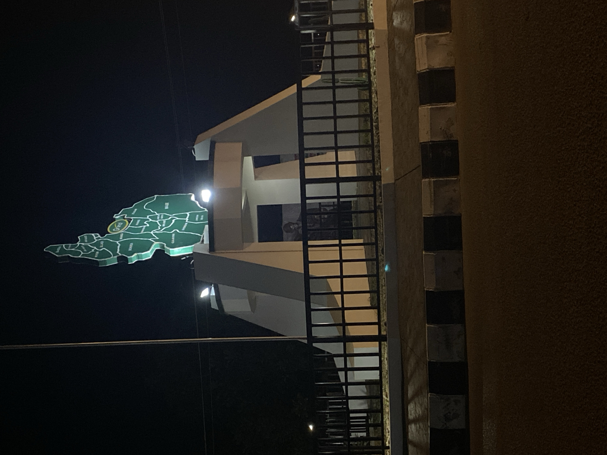 This nighttime image, captured in Bukuru, Nigeria, showcases an illuminated monument featuring a large, prominently displayed map of Nigeria. The map is green with white outlines delineating the states and is mounted on a modern architectural structure with angular white and beige surfaces.
Numerous Nigerian states are clearly labeled on the map, including "KADAM", "BAUCHI", "GOMBE", "TARABA", "BENUE", "PLATEAU", "NASARAWA", "KANO", "JIGAWA", "KATSINA", "ZAMFARA", "SOKOTO", "KEBBI", "NIGER", "KOGI", "FCT", "OYO", "OSUN", "ONDO", "EKITI", "OGUN", "LAGOS", "ANAMBRA", "EBONYI", "IMO", "ABIA", "RIVERS", "BAYELSA", "DELTA", "EDO", "AKWA IBOM", "CROSS RIVER", "ADAMAWA", "BORNO", and "YOBE". A "Grand Park" logo is also visible on the map.
The structure itself is lit by several artificial lights, highlighting its features and the map against the dark sky. To the right, a black metal railing extends vertically, likely part of a staircase or an elevated walkway. Further right, a wall with distinct black and white striped blocks or patterns is partially visible. The scene is devoid of people or activity, focusing solely on the illuminated monument.