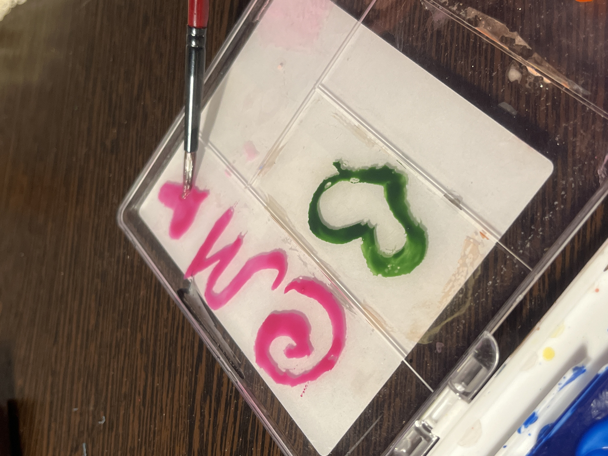 A top-down, close-up shot depicts a clear plastic paint palette resting on a brown, wood-grain surface. The palette is divided into compartments. Within the compartments are pink and green paint. A paintbrush with a red handle rests in one compartment with pink paint. The pink paint is also arranged as the letter "w" and a spiral design. The green paint is arranged in a heart shape. A portion of another paint palette is visible in the lower right corner containing white, yellow, and blue paint. The location is not discernable from this image.