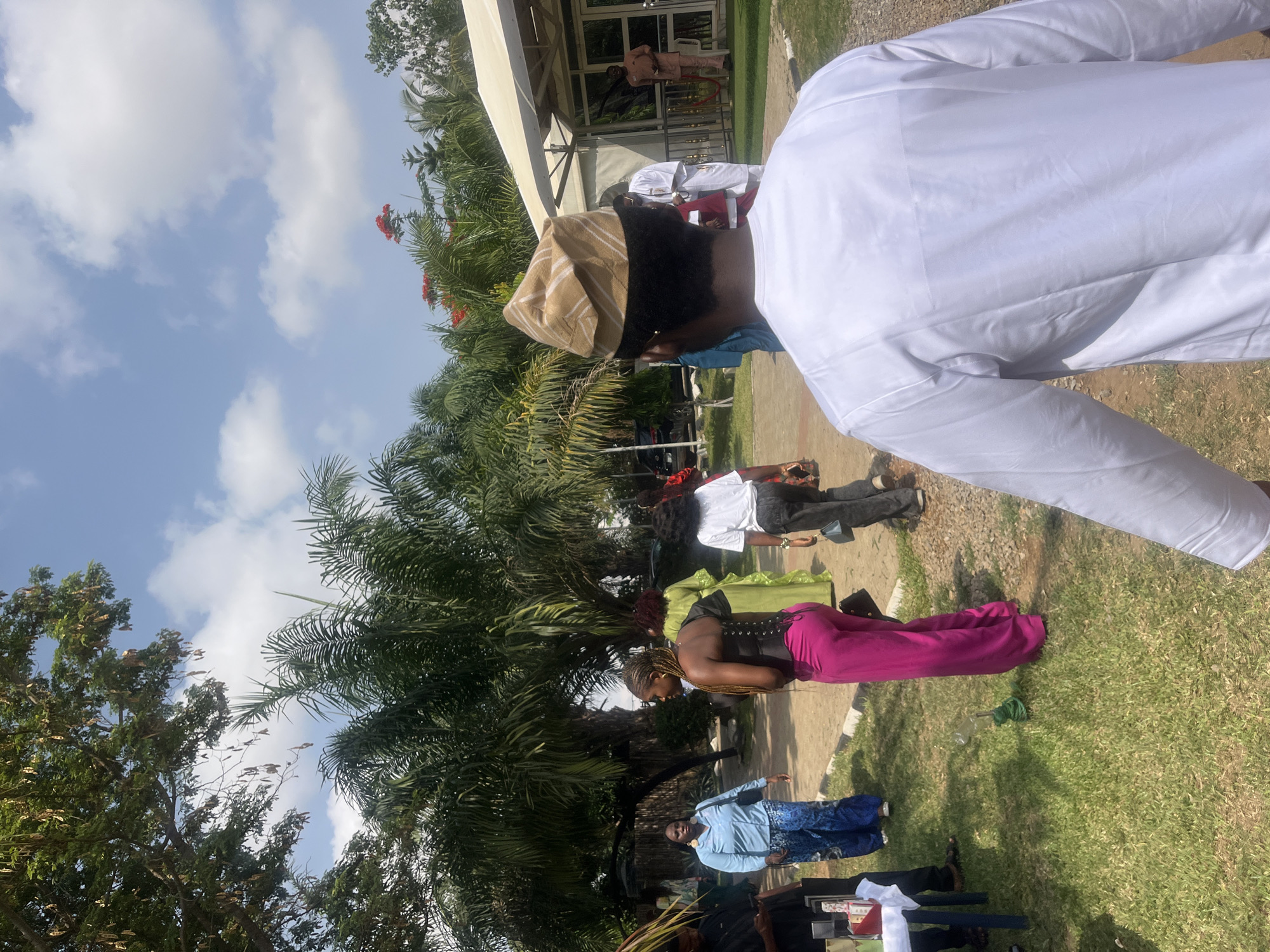 A ground-level, eye-level outdoor shot depicts a gathering of people at Galadimawa Village, Nigeria. The sky is blue with scattered clouds. A person in a white shirt stands in the foreground. Several other people are present, including one wearing pink pants and a black top, and another in a blue outfit. Some individuals are partially obscured. Green grass covers the ground, and a building structure is visible in the background behind the people. Palm trees and other foliage are present, adding to the outdoor setting.