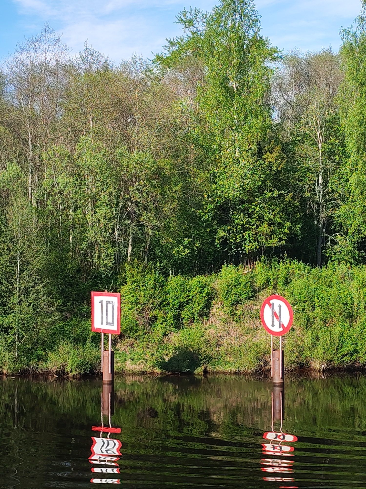 The image depicts a waterway in Devyatiny, Russia, flanked by dense green foliage and trees. Two navigational markers are visible at the water's edge. The marker on the left is a square sign with the number "10" displayed. The marker on the right is a circular sign with a red border and a white center featuring a vertical line with opposing arrows, indicating a restriction or prohibition of some kind. Both markers are reflected in the dark, rippling water. The sky above is light blue with wisps of clouds.