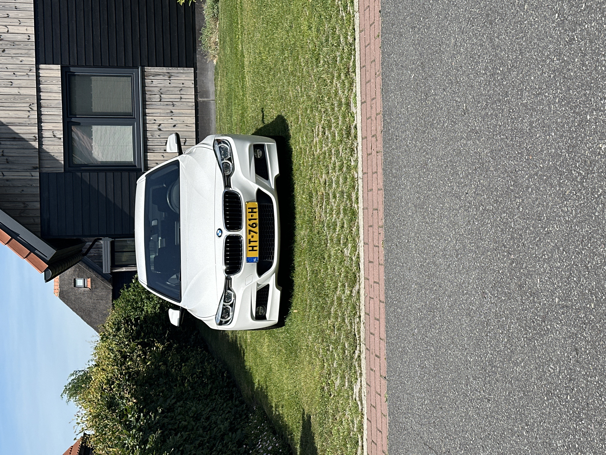 In Ossenzijl, Netherlands, a white BMW sedan is parked on a grassy driveway bordered by a red brick walkway and asphalt road. The car faces the viewer, with its license plate reading "HT-761-H". Behind the car, a dark-colored modern house with a wooden facade and a thatched roof is visible. A neatly trimmed green hedge lines the left side of the driveway. The sky is clear and blue.