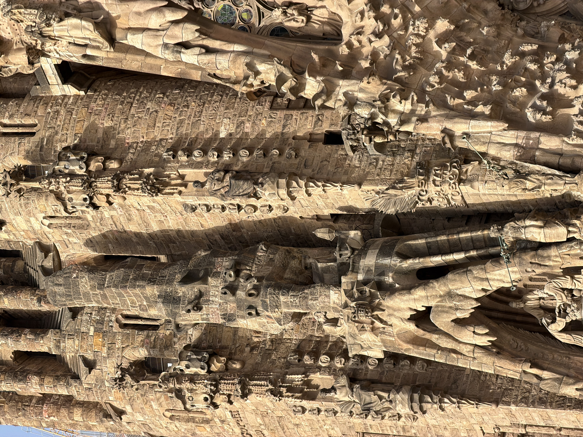 The image shows a detailed close-up of the facade of the Sagrada Familia in Barcelona, Spain. The architecture is characterized by intricate stonework, featuring textured walls, columns, arched openings, and numerous sculpted figures and decorative elements. Sunlight casts shadows, highlighting the three-dimensional aspects of the carvings. The stone material appears to be a warm, sandy-brown color. Several statues are visible, depicting human figures in robes, integrated into the architectural design. The scale suggests a monumental structure, with rich ornamentation in a Gothic-inspired style.