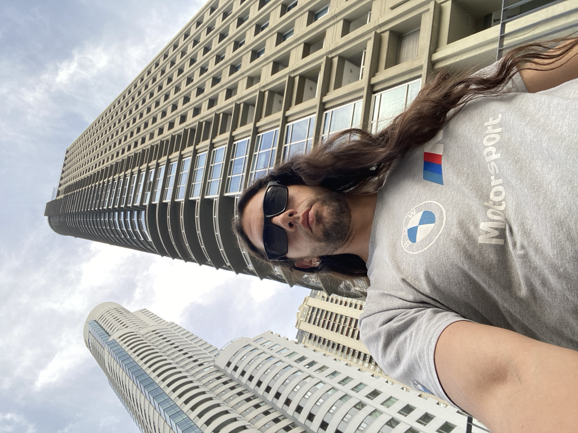 A man with long brown hair, wearing sunglasses and a grey BMW Motorsport t-shirt, is taking a selfie in front of two tall buildings in Buenos Aires, Argentina. The building directly behind the man is a tall, beige building with a grid-like pattern of windows and balconies, characteristic of rationalist architecture. To the left and slightly behind this building is a second, white, curvilinear tower. The sky is overcast with grey clouds. The photograph is taken from a low angle, emphasizing the height of the buildings.
