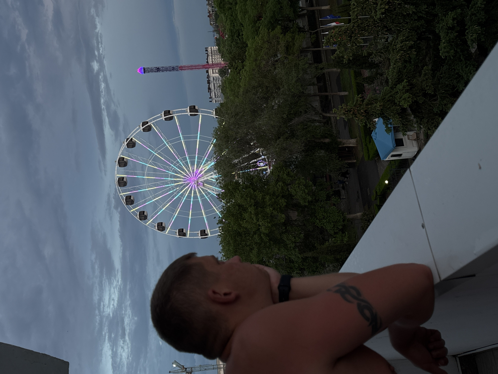 The image shows a man standing on a balcony overlooking a Ferris wheel and a tower attraction. The Ferris wheel is brightly lit with various colors. Trees and buildings are visible in the background, along with an overcast sky. Based on the landmarks, the location is Yevpatoriya, Ukraine. The man is bare-chested, wearing a wrist-worn device, and has a tattoo on his upper arm. He appears to be looking at the Ferris wheel. A white structure, likely part of the balcony, is in the foreground.