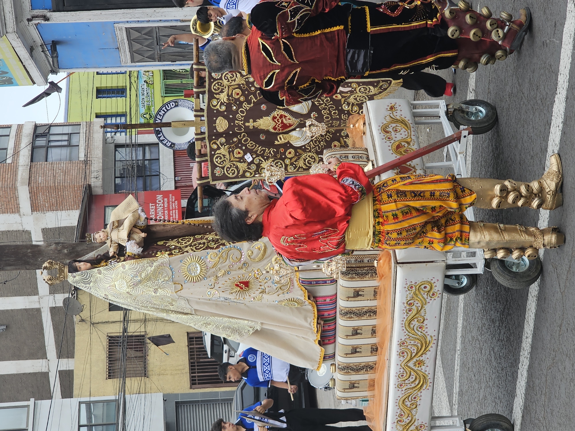 The image shows a religious procession in Iquique, Chile. A large statue of the Virgin Mary with the child Jesus is being carried on a decorated float. The float is adorned with intricate gold designs and a blanket featuring llamas. Several people are participating in the procession. Two young men in blue and white uniforms are playing instruments, one with cymbals and the other with a drumstick. A man in a red and yellow outfit, with gold boots adorned with bells, is holding a pole. Another man in a dark red and black outfit, also with bell-adorned boots, stands to the right. In the background, buildings with colorful facades and power lines are visible, indicating an urban setting. A banner with Spanish text, possibly related to the religious event, is also present. The overall scene suggests a vibrant cultural or religious festival.