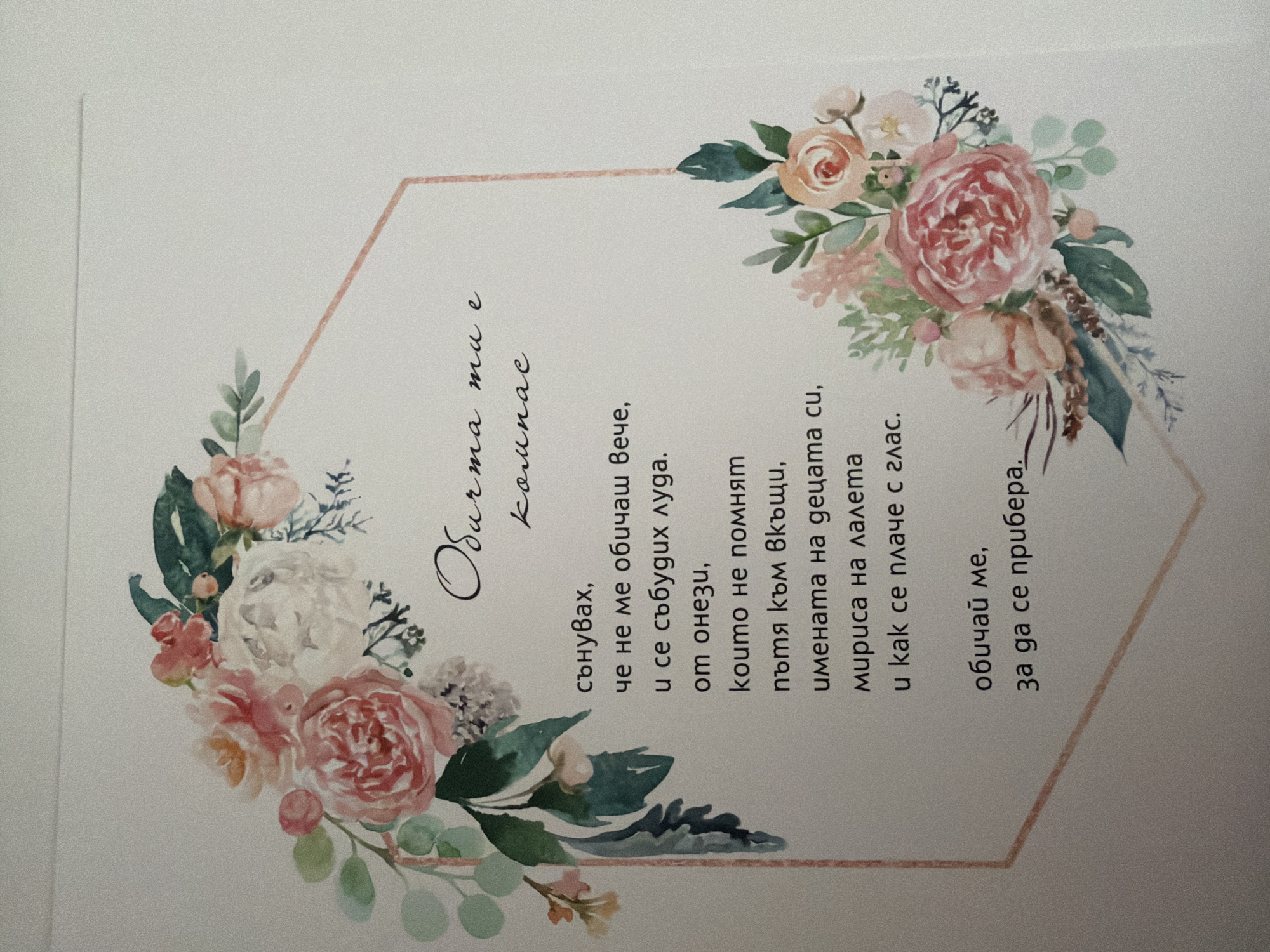 The image is a close-up, high-angle shot of a greeting card. The card features a floral watercolor design with pink and white roses and green foliage framing a hexagonal shape. Inside the hexagon, there is text in Bulgarian. The top line of text is in a stylized script and reads "Обичам те" (I love you). Below this, in a more standard font, it says "стаи" (rooms), although the final letter might be a lowercase "e" to form "стая" (room). The following lines of text are in a poem format, listing dreams, waking up, forgetfulness, paths, names, scents, and a voice. The card is placed on a white or very light-colored surface, possibly a table or wall. There are no people, actions, or specific environmental details of Pazardzhik, Bulgaria visible in the image.
