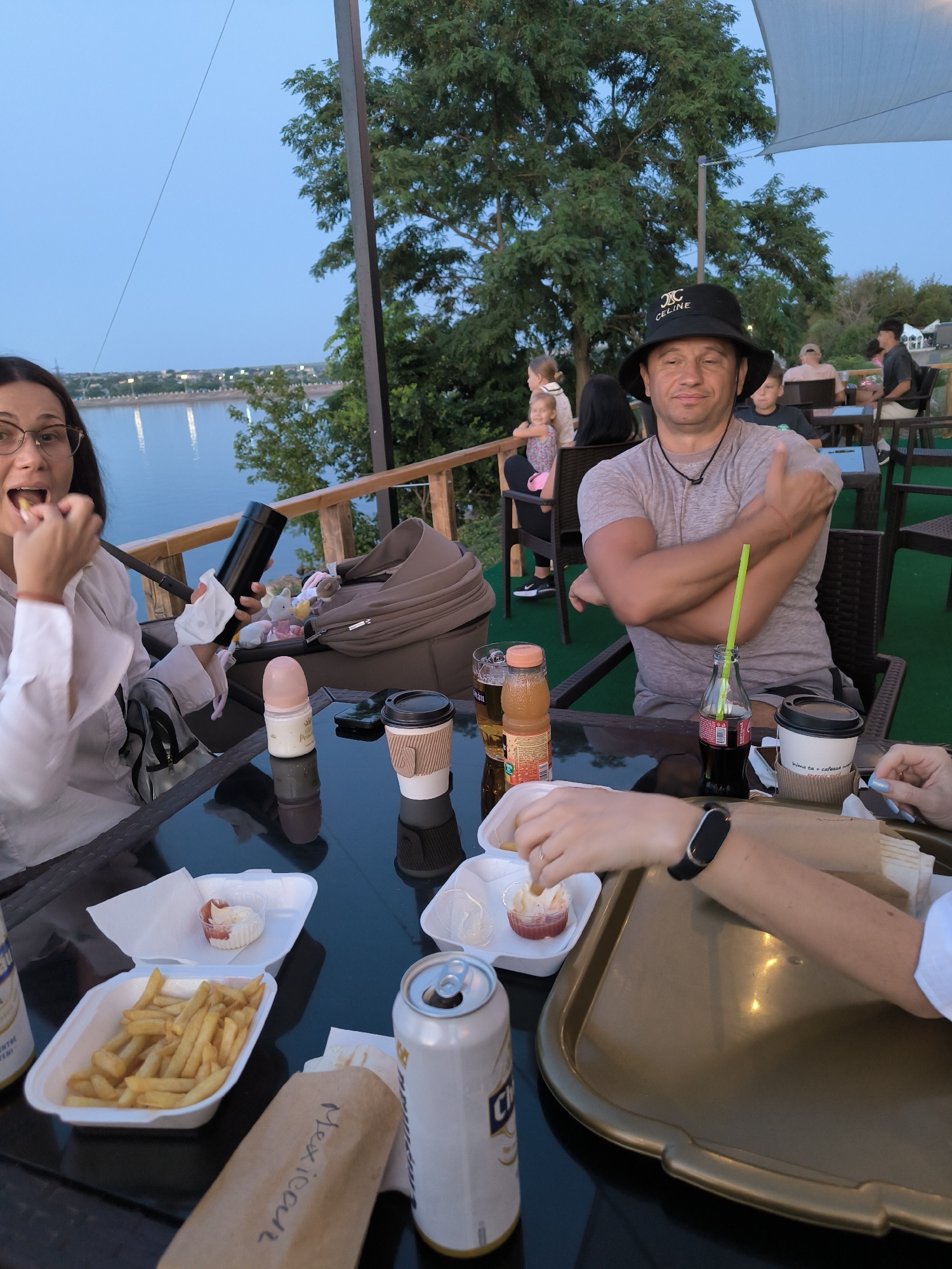 The image depicts a group of people gathered at an outdoor dining area near a body of water, possibly in Ustia, Moldova. The setting appears to be a restaurant or cafe with tables and chairs on a deck overlooking a river or lake. The time of day is dusk, with a twilight sky.
In the foreground, a woman wearing glasses and a white shirt is seated at a table, eating. She is holding a piece of food in her right hand and a black water bottle in her left. Beside her, on the table, are various items including a white tissue, a baby bottle, a black cup with a lid, and two open containers of food - one with french fries and another with a small portion of dipping sauce. A can of soda and a bottle of juice are also visible on the table.
To the right of the woman, a man wearing a black bucket hat and a grey t-shirt is seated with his arms crossed. He is giving a thumbs-up gesture. He is also seated at the table with drinks and food. A green straw is visible in a dark colored soda bottle.
In the background, other patrons are seated at tables. A stroller with stuffed toys is placed behind the woman in the foreground. Trees and foliage surround the dining area, and a railing is visible along the edge of the deck. The overall scene suggests a casual gathering or meal.