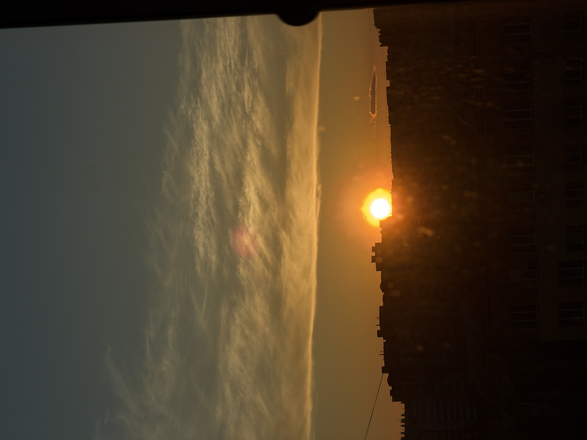 The image depicts a sunset view in the city of Krasnoyarsk, Russia. The sun is positioned on the horizon, partially obscured by the silhouetted skyline of a tall building. The sky is filled with wispy clouds, illuminated by the setting sun in shades of yellow and orange. The building appears to be a residential structure, with a grid of windows visible in the dark facade. A power line can be seen extending from the buildings towards the bottom of the frame. The overall image is backlit, creating strong contrasts and a sense of twilight.