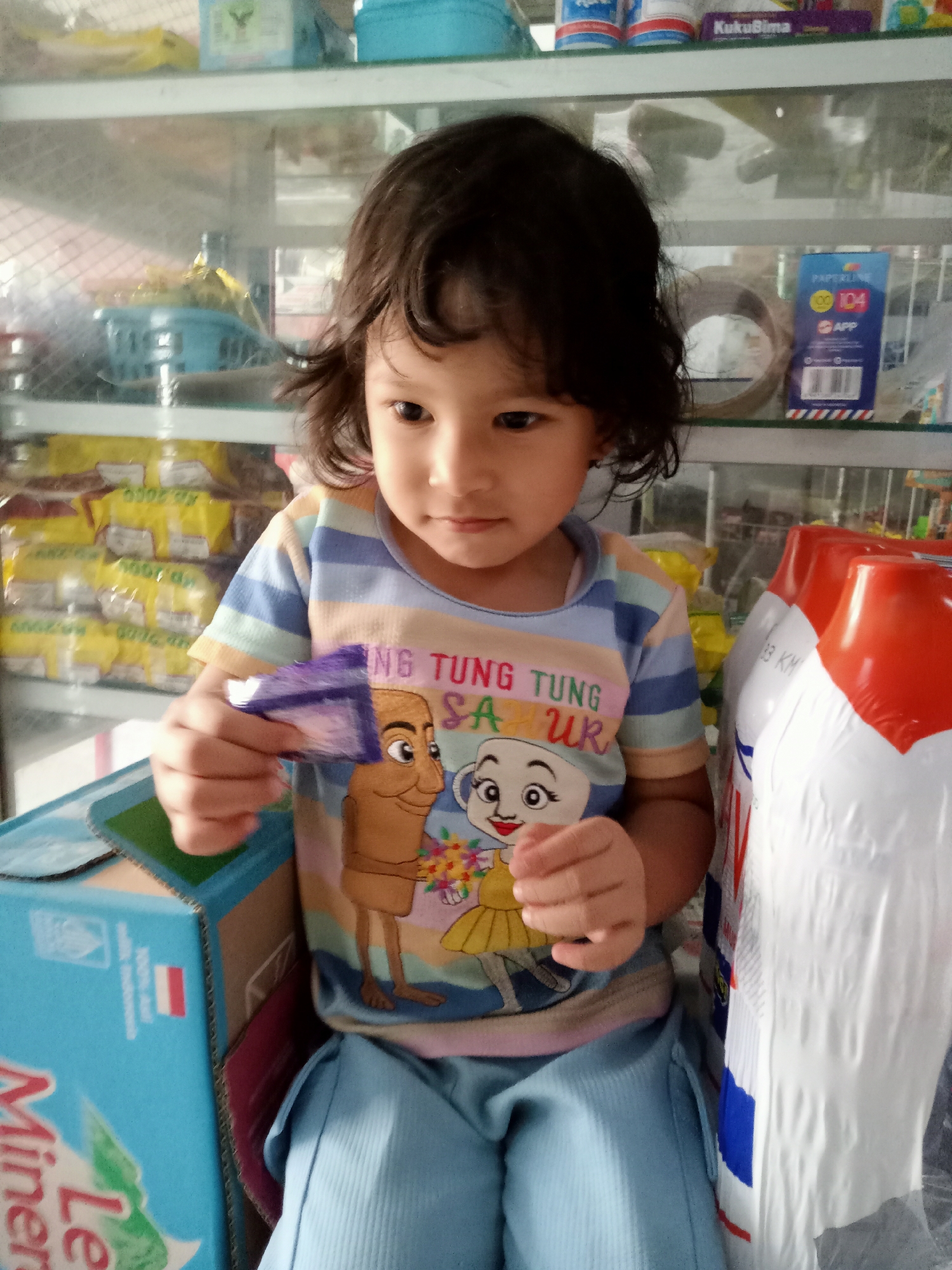 In Simo, Indonesia, a young girl with dark, curly hair is holding a small, purple, wrapped item in her hands. She is wearing a striped t-shirt with cartoon characters on it, and light blue pants. The background shows shelves stocked with various products, including what appear to be bags of snacks and bottles of cleaning supplies. The scene suggests a retail environment, possibly a small shop or market stall.