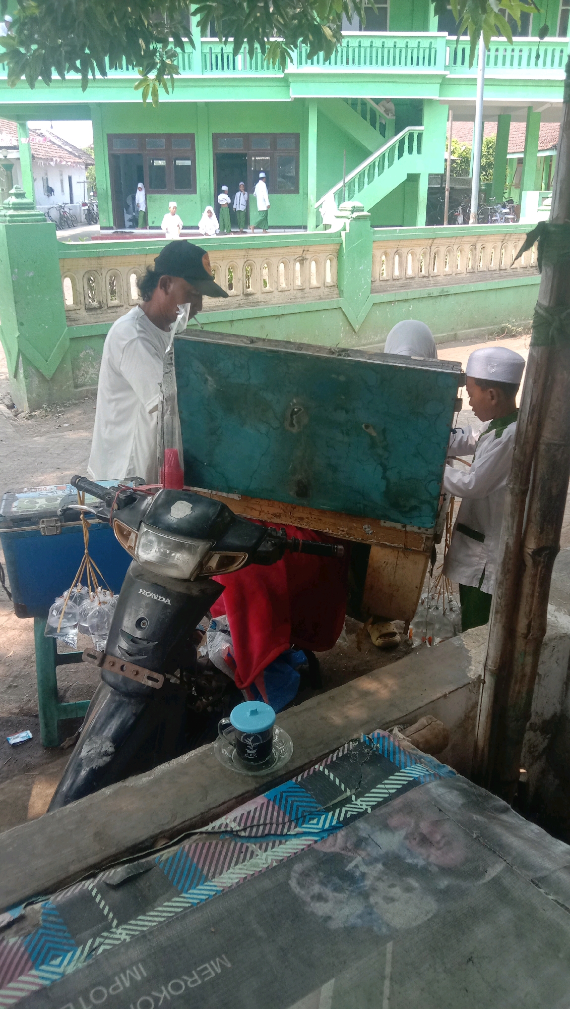 In front of a green building, possibly a school or mosque, in Ngempit, Indonesia, a man wearing a white t-shirt and a black baseball cap stands by a scooter. He appears to be attending to a mobile beverage cart. The cart, constructed from wood and metal, is topped with a large, turquoise-colored box. Below the box, a red cloth is visible. The scooter is equipped with a cooler box on the left side, from which hang several small plastic bags containing liquid. A young boy in a white tunic and cap is interacting with the cart. In the foreground, a table is covered with a patterned cloth, and on it sits a small cup of dark liquid with a blue lid and a saucer. The environment suggests an outdoor setting with a paved or dirt ground.