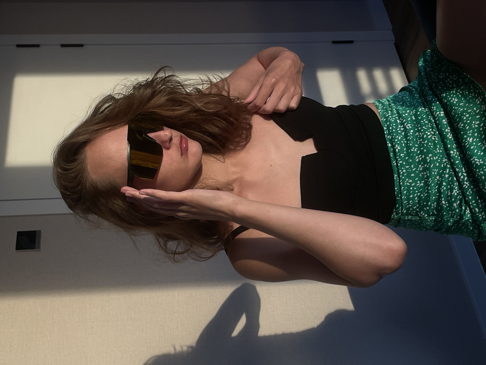 The image features a woman wearing sunglasses, a black top, and a green floral skirt. She is reclining with one hand near her face. The background consists of a wall and door with sunlight casting shadows. There is no indication of the location being Подшивалово, Russia.
