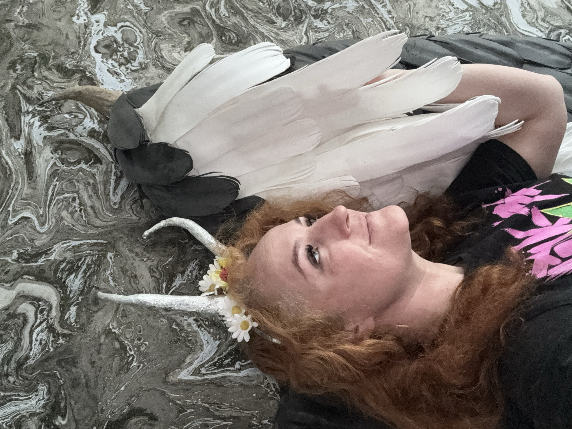The image depicts a woman lying on a surface with a swirling grey and white pattern. She is facing upward and has red curly hair, silver horns adorned with daisies, and is wearing a black shirt with pink designs. Next to her is a costume piece resembling a bird's head, featuring a brown beak, black head covering, and white feathers. The scene does not show anything that confirms that it is in Chaikovsky, Russia.
