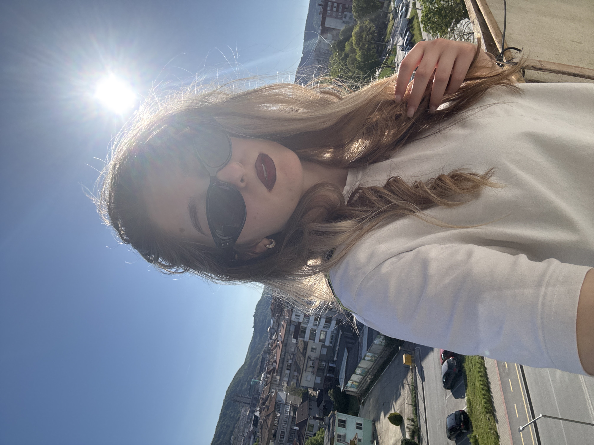 A woman is taking a selfie with a city in the background. The city appears to be nestled between a hill/mountain and a road with multiple cars, potentially in the city of Unknown, Unknown. The woman is wearing sunglasses and dark lipstick, and she has her hand in her hair. The sun is shining brightly, creating a lens flare effect in the upper left corner of the image.