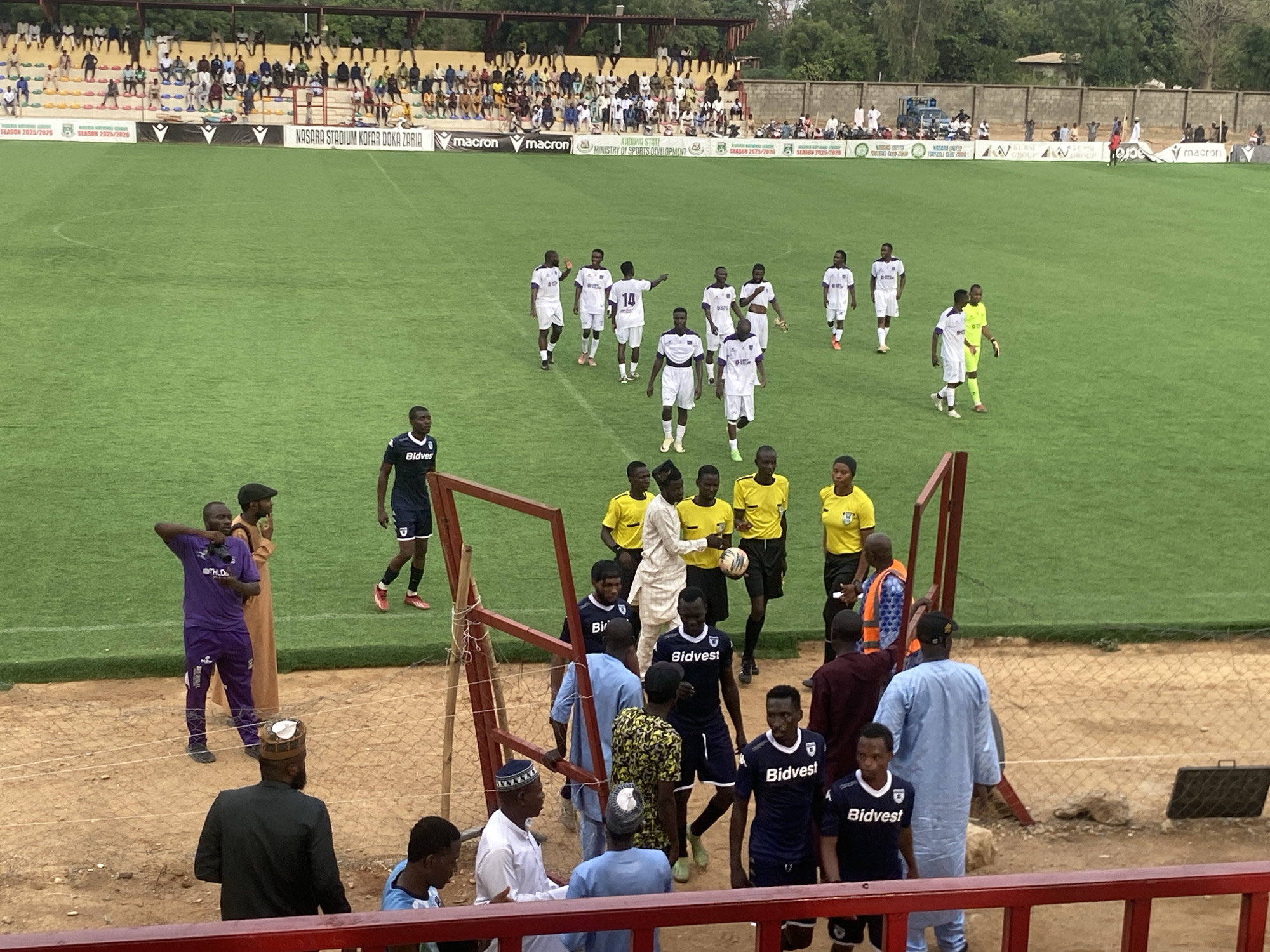 The image captures a scene at Nasara Stadium Kofar Doka Zaria, Tudun Wada, Nigeria, likely during or after a soccer match. On the green pitch, players in white and purple uniforms are walking towards the stands. Near the boundary line, a group of people, including players in navy blue uniforms with "Bidvest" printed on them, referees in yellow shirts, and other individuals in casual clothing, are gathered near an opening in the fence. The stands are filled with spectators.