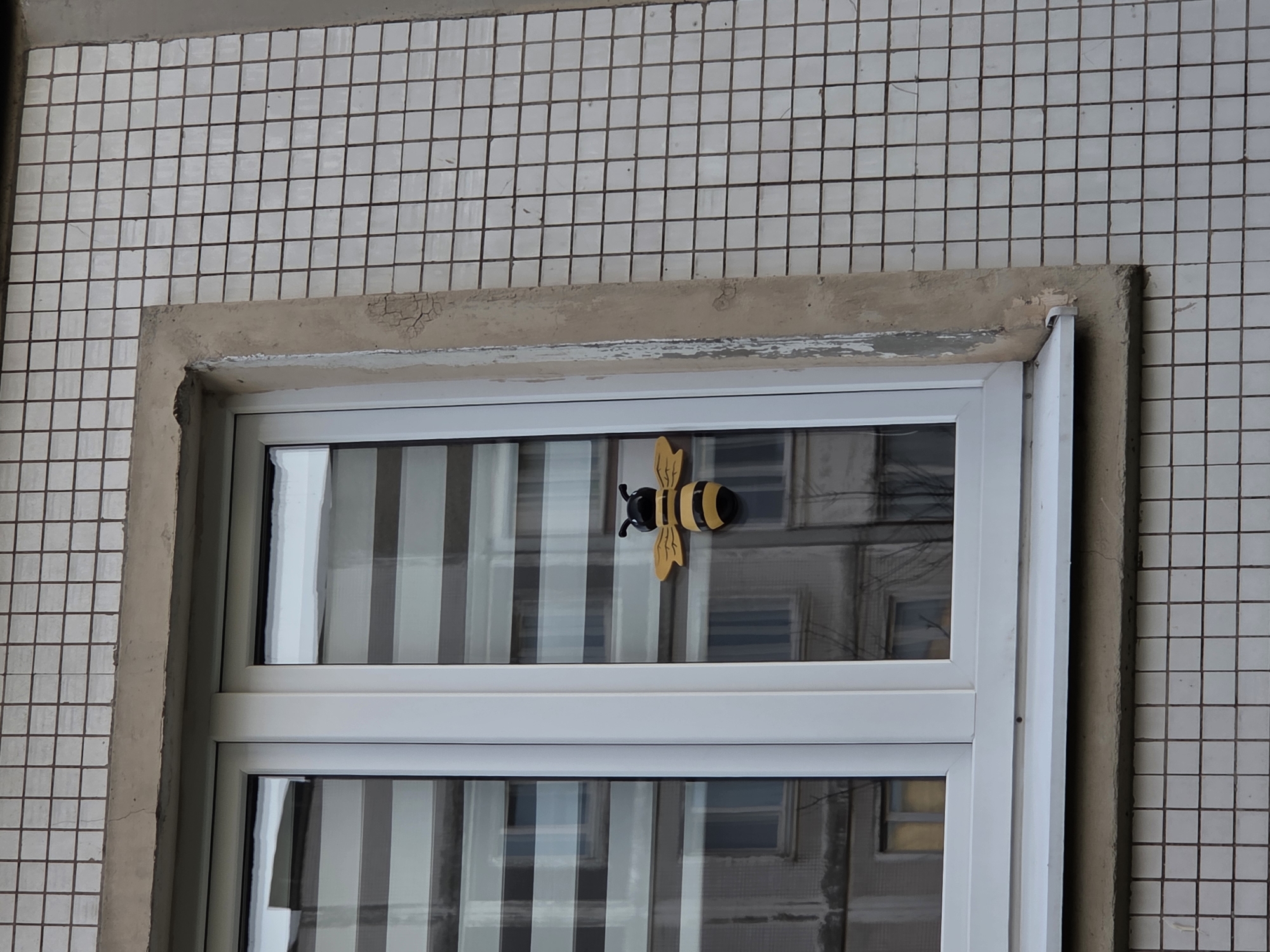 Here is a forensic description of the image:
**Scene:** A window is set into a tiled wall. The tiles are small, white, and square, with dark grout lines. The window has a heavy, weathered concrete frame. The window itself is white and appears to have two panes.
**Subject:** A decorative bee is affixed to the outside of the window. It is black and yellow, with yellow wings.
**Environment:** The window reflects an image of another building with windows, suggesting an urban environment. The city and region are unknown.
**Objects:** Besides the bee, window, and tiled wall, there are no other immediately discernible objects.