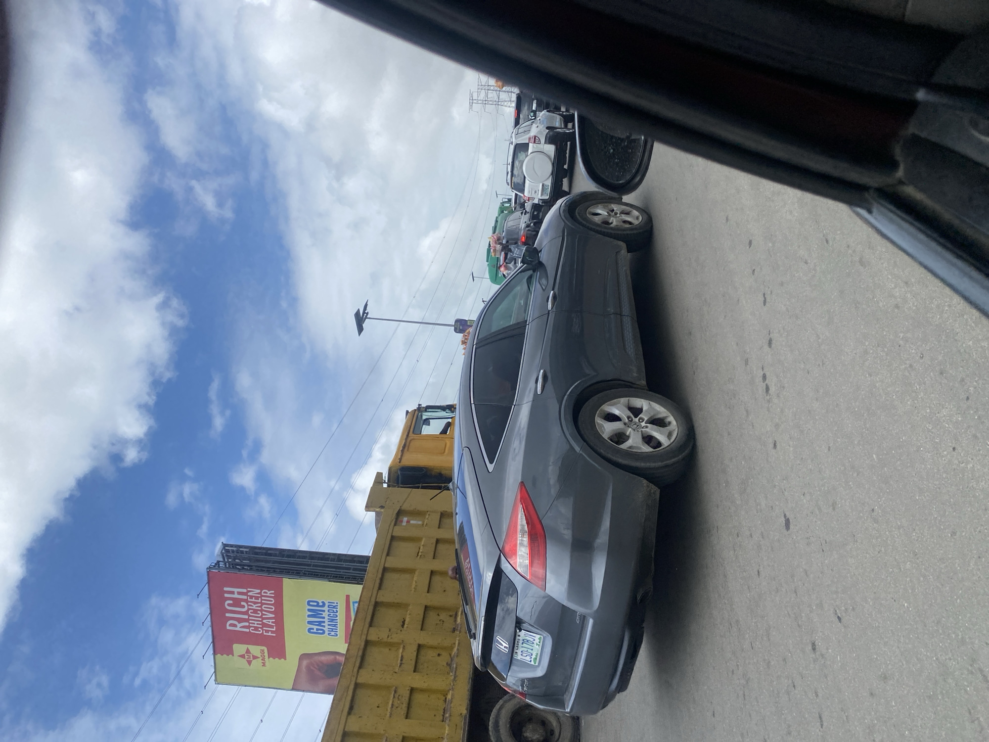 In Jakande, Nigeria, a grey Honda Cross tour is seen in traffic on a road under a cloudy sky. The car's license plate reads "LSD-178-JV". In the background, a large yellow dump truck is parked next to a billboard that advertises "RICH CHICKEN FLAVOUR" and "GAME CHANGER!" by Maggi. Other vehicles, including SUVs and a green truck carrying goods, are also visible in the congested traffic. Power lines and streetlights are present in the urban environment.