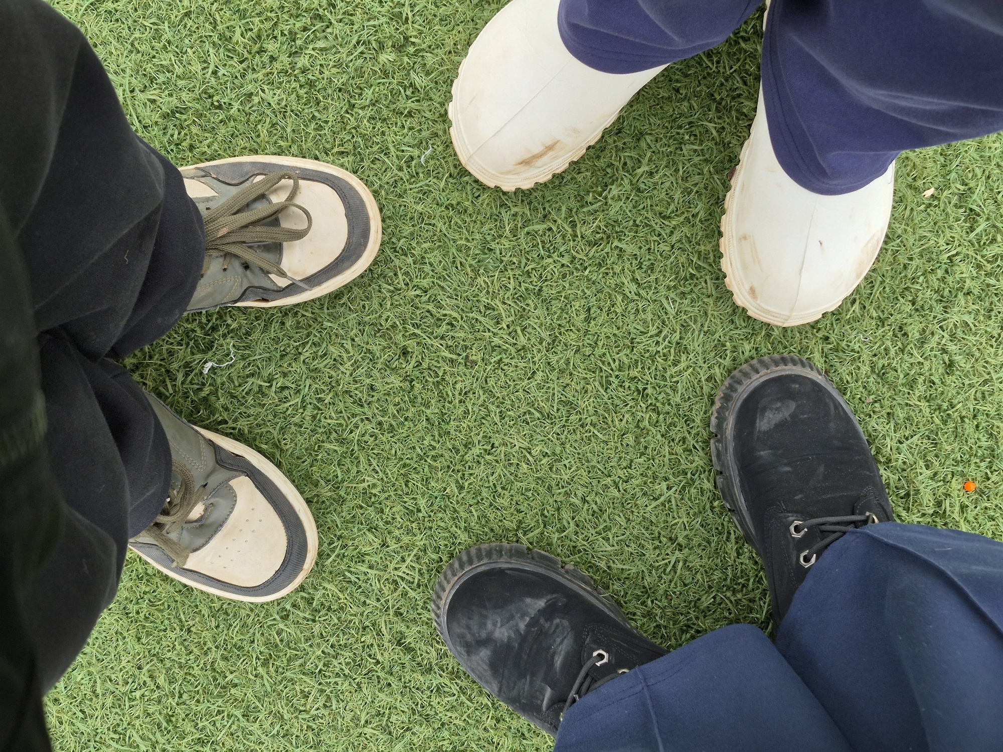 Three pairs of feet are on a green, artificial turf surface. The pair on the left wears sneakers with off-white soles and green laces and uppers with black pants. The pair at the top wears white rubber boots with navy blue pants. The pair on the right wears black work boots with navy blue pants. There is a small, orange circular object in the lower right corner. The location is unknown, but the city of Chaikovsky, Russia, cannot be verified from the image.