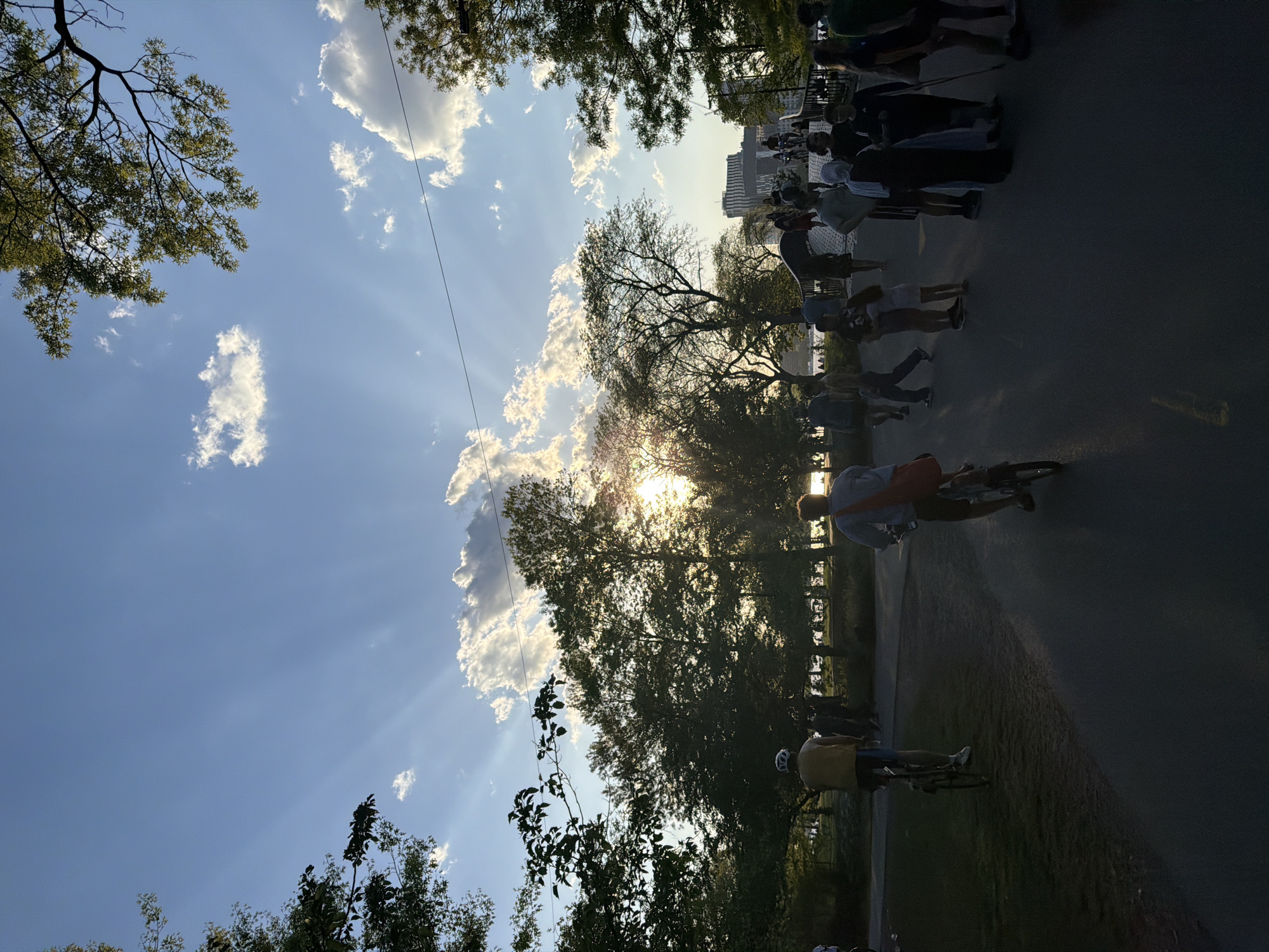 This image depicts a sunny day in Boston, Massachusetts, most likely in the Back Bay Fens or on the Charles River Esplanade, as indicated by the visible skyline and trees. Several people are walking and riding bikes on a path, suggesting a park-like setting. The sky is predominantly blue with scattered clouds, and the sun is positioned behind the trees, casting rays of light. The people appear to be engaged in recreational activities. A group of buildings is visible in the background. A cyclist is traveling on the path.