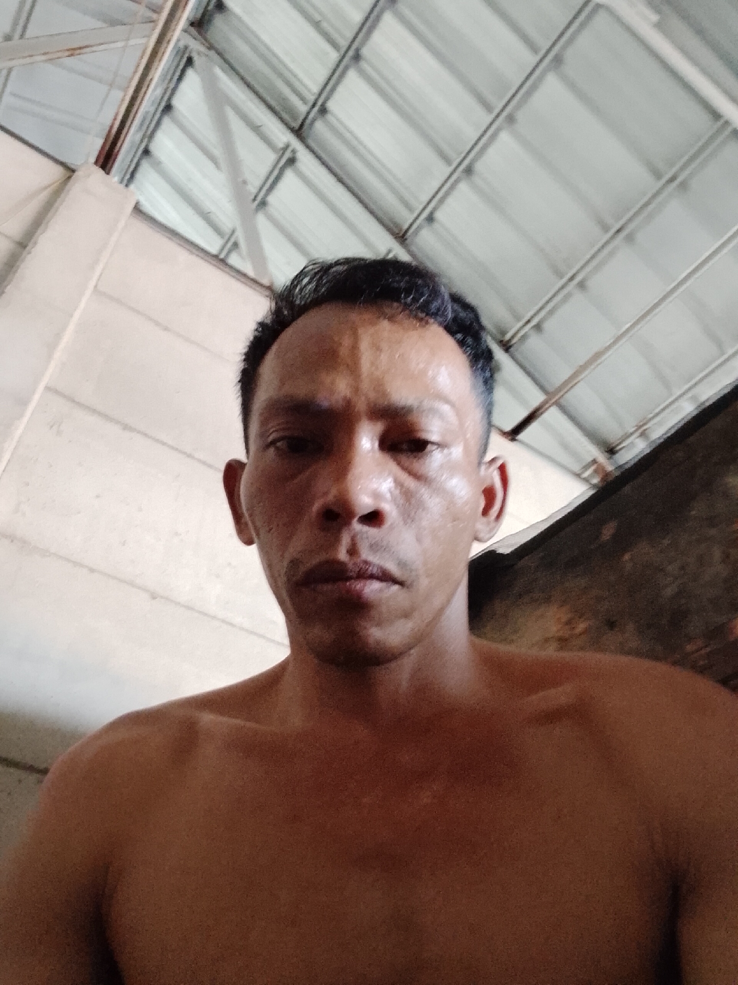 A medium close-up angled shot shows a shirtless man in Bukit Ketok, Indonesia, with short dark hair. He has dark skin and is looking directly at the camera. He appears to be indoors in a space with a metallic corrugated roof and white walls.