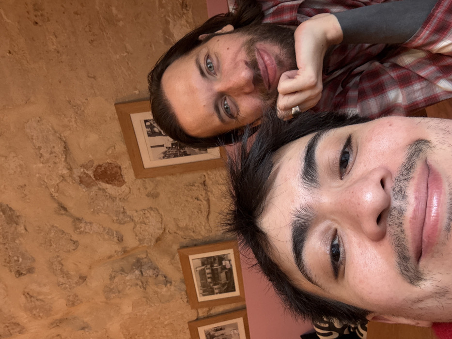 The image displays a close-up selfie of two individuals, both appearing male, inside a building in Paris, France. The subject in the immediate foreground has dark hair, prominent eyebrows, and a light mustache, looking directly at the camera with a slight smile. Partially visible on the right is a second subject with long dark hair, a beard, and light-colored eyes, also smiling while resting his chin on his right hand, which wears a silver ring. He is dressed in a red and white plaid shirt over a dark long-sleeved garment. The background consists of an exposed, irregular stone wall characteristic of historic Parisian architecture. Two framed black and white photographs depicting urban street scenes are mounted on this wall. A section of a pink or mauve upholstered surface is visible behind the foreground subject's head. The setting suggests a casual indoor environment, such as a café or restaurant in a historic district of Paris.