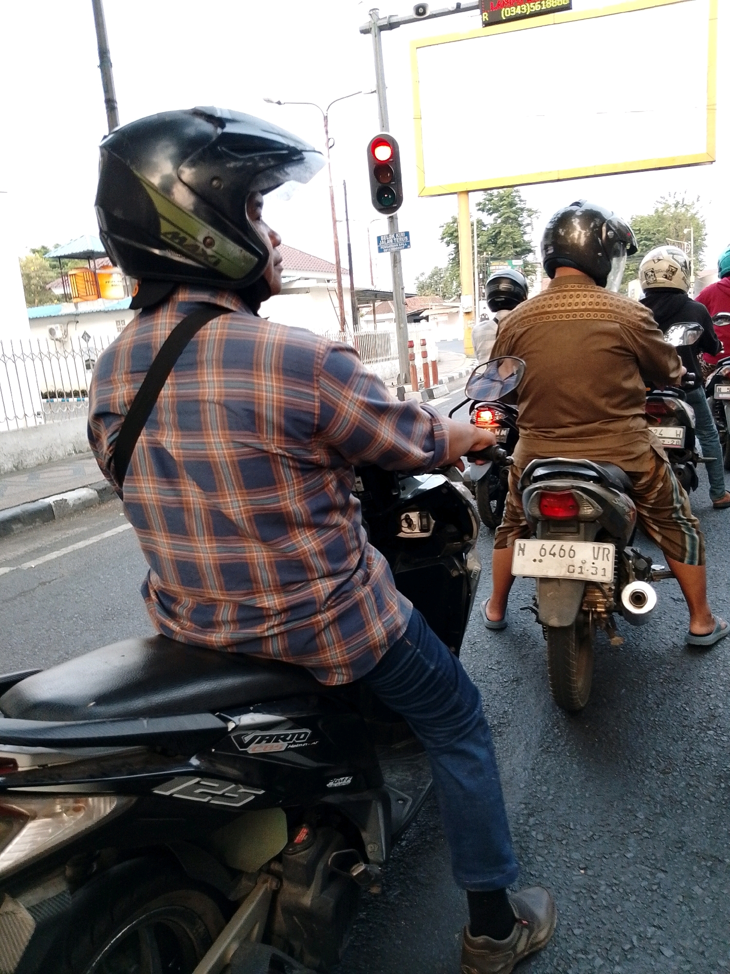 The image, taken in the city of Pasuruan, Indonesia, depicts a street scene with multiple motorcycles and riders. In the foreground, a person wearing a plaid shirt and blue jeans is riding a black scooter, identified by the "VARIO 125" logo on the side. The rider is wearing a black helmet with green accents. Behind this scooter, another rider wearing a brown patterned shirt and striped pants is on a motorcycle. This motorcycle has a license plate that reads "N 6466 UR" and "01-31". Further back, another motorcycle with a rider in a helmet is visible, and parts of a fourth motorcycle and rider are also present. The background includes a traffic light displaying red, a billboard, trees, buildings, and a white fence. The road surface is asphalt.