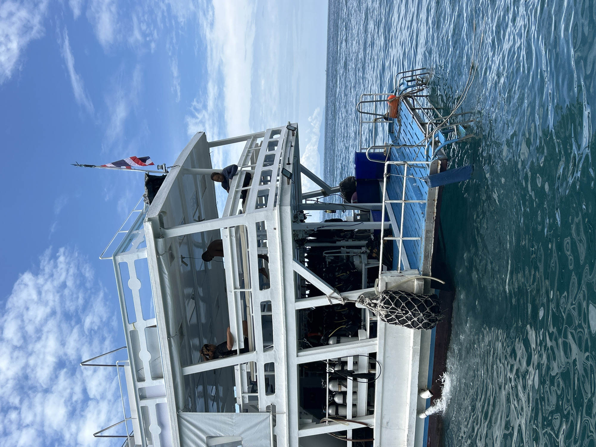 An external, slightly low-angle, eye-level shot captures the stern of a white dive boat on a calm, blue ocean under a bright, cloudy sky. The boat's blue deck is visible at the stern, with a metal ladder extending into the water. An orange life preserver is attached to the railing. A Thai flag flies from a mast on the upper deck, where two individuals are seated on the right. The boat's hull has a blue waterline. The lower deck of the boat is packed with scuba diving gear, including numerous air tanks. A black fender hangs from the side of the boat, and water is seen splashing from a pipe on the hull. The background shows a distant, hazy coastline. The scene is set in Ao Nang, Thailand.