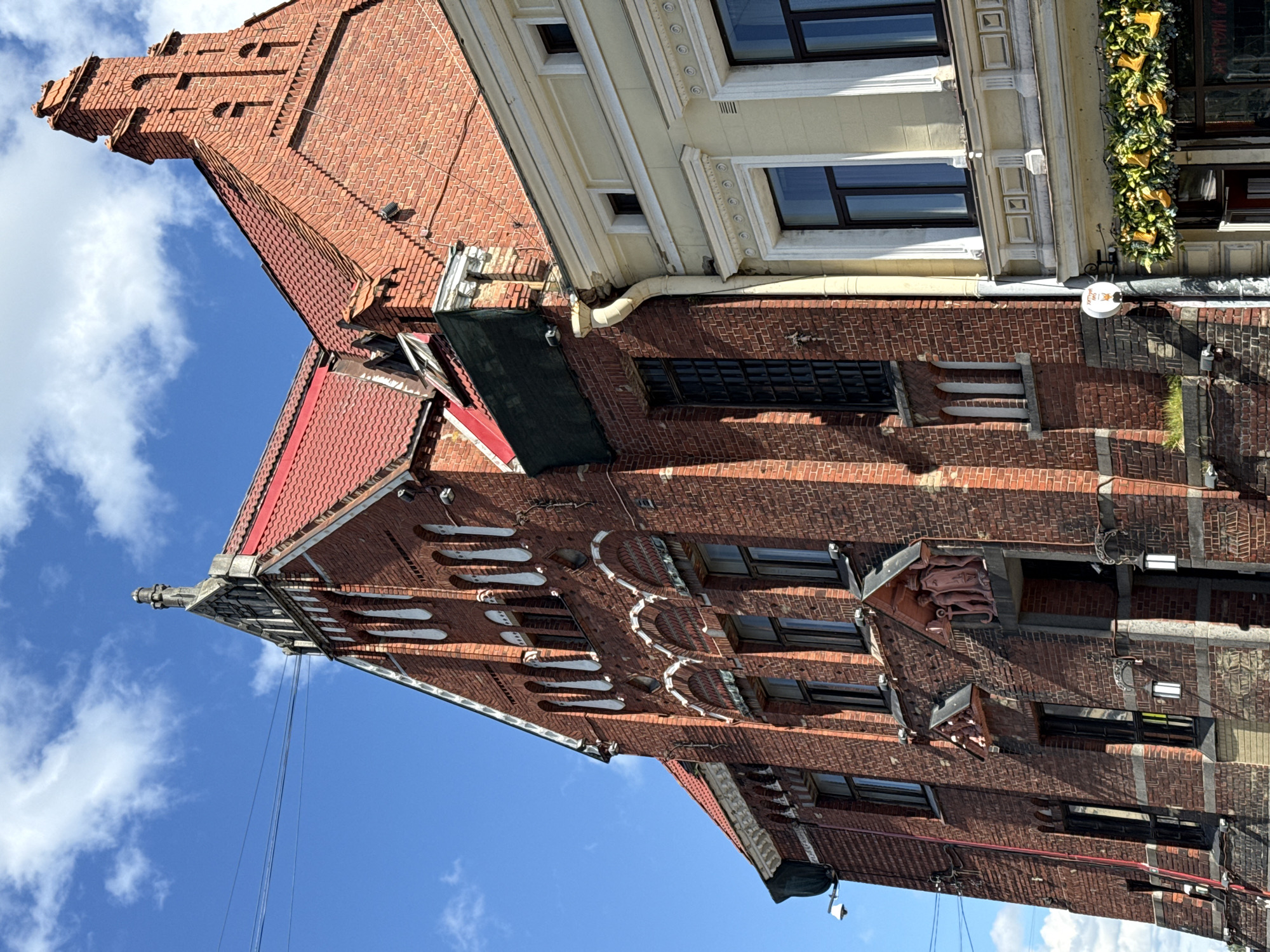 The image shows the exterior of a brick building with a red tile roof in Vyborg, Russia. The building is multi-story and features arched windows and decorative white trim. To the right of the brick building, there is a lighter-colored building with a similar architectural style. Both buildings are situated under a bright blue sky with scattered white clouds. The scene appears to be a street view, with power lines visible in the background. The overall impression is one of historical architecture in an urban setting.