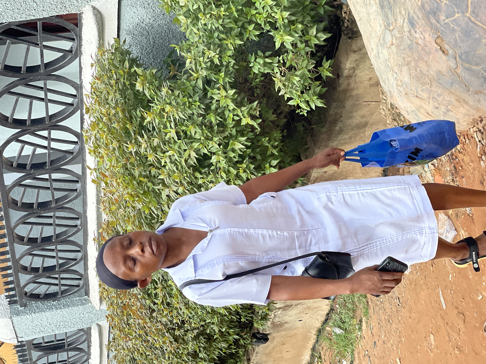 A woman stands on a dirt and paved surface, likely a sidewalk or roadside in Nkpor, Nigeria. She is wearing a white dress and sandals, carrying a blue shopping bag in her left hand and a black smartphone in her right. A small black purse hangs from her shoulder. She is standing in front of a building with a distinctive window design (circular with horizontal bars). A bush with green leaves is behind her. The image appears to be taken outdoors in daylight.