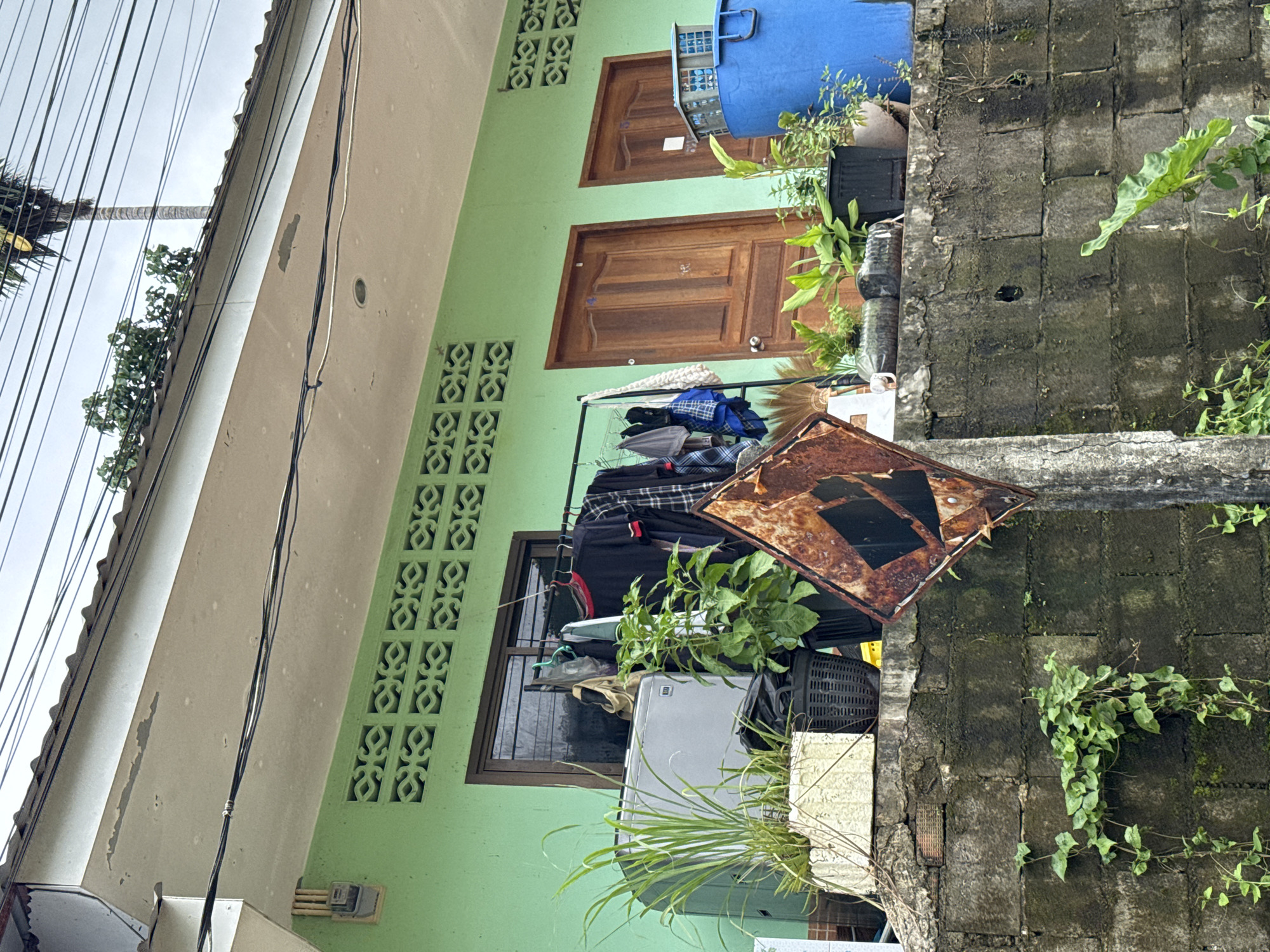 A scene in Rawai, Thailand, features a light green stucco building with two wooden doors and a large window. Clothes hang on a rack in front of the window, and a silver washing machine sits to the left of the rack. Several potted plants are arranged on a low concrete wall in front of the building, some with lush green foliage. A rusted diamond-shaped signpost stands near the wall. A large blue plastic barrel and a blue basket are visible on the right side of the frame, near the doors. The sky is overcast, and power lines run across the top of the image. Overgrown vegetation climbs the concrete wall.