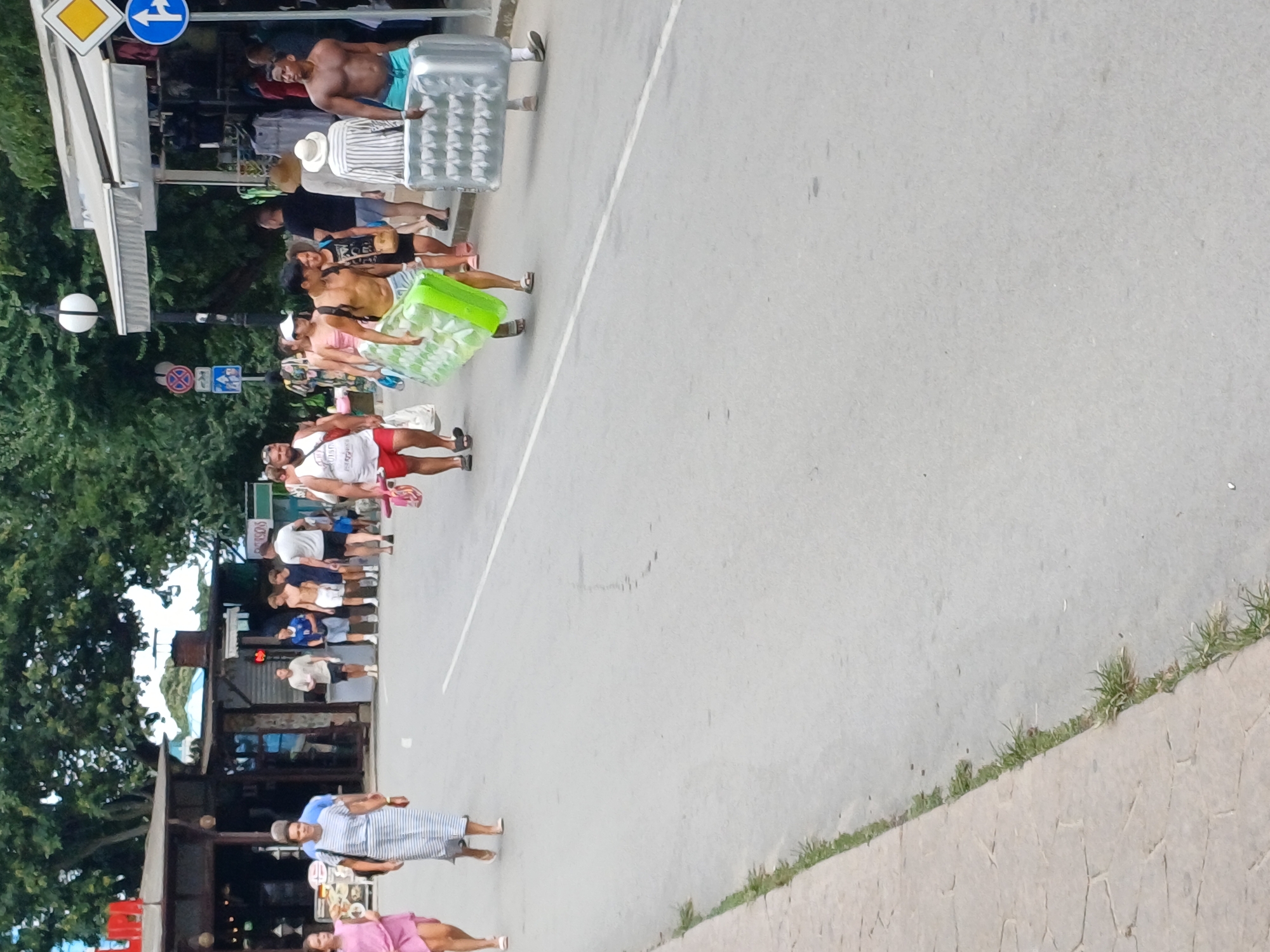 A group of people are walking on a street, likely in Varna, Bulgaria, given the context of the location. In the foreground, two individuals are carrying large inflatable mattresses. The person on the left is a man with dark skin and short hair, wearing blue swim trunks and carrying a bright green inflatable. The person on the right is wearing a striped shirt and a hat, and is carrying a silver inflatable. Behind them, a woman in a white and blue striped dress is walking, and further back, a woman in a pink dress is also walking. There are shops and stalls lining the street on the right side, with awnings providing shade. Trees with lush green foliage are visible in the background. Several other people are walking in the middle ground, some of whom appear to be tourists. The street is paved with asphalt, and there is a sidewalk with some grass growing along its edge on the left side of the image. Traffic signs, including directional arrows and a yellow diamond shape, are mounted on poles along the side of the street. The overall scene depicts a bustling street environment, possibly near a beach or resort area.