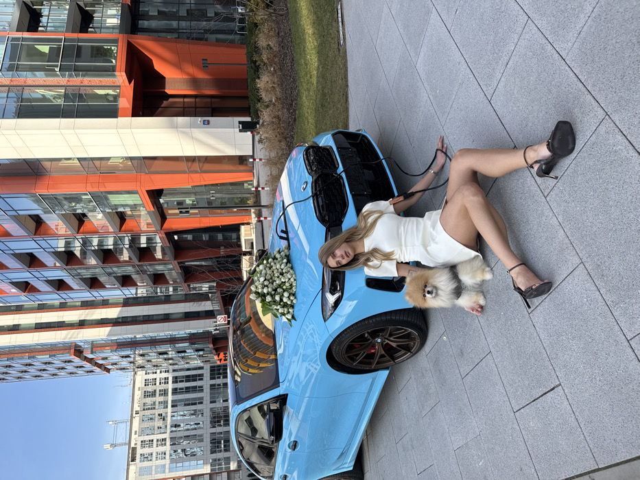 A light blue BMW 3 Series sedan is parked on a grey paver pathway within the modern urban development of Browary Warszawskie in the Wola district of Warsaw, Poland. The vehicle features a black grille, black roof, and bronze-colored multi-spoke alloy wheels. A large bouquet of white roses is positioned on the car's hood, and two black cables extend from the front of the car towards the ground and a seated woman.
A young adult female with long light brown hair is seated on the pavement to the front-left of the car. She wears a white short-sleeved mini-dress and black high-heeled open-toed shoes with ankle straps. A black clip from one of the cables is attached to her right forearm, while another cable runs from the car towards her left side. She looks directly at the viewer with a neutral expression.
To her immediate left, a small, fluffy Pomeranian dog with light brown fur and a grey harness sits on the pavement, also looking towards the viewer. The woman's left hand rests near the dog.
In the background, modern high-rise buildings with glass facades and distinctive orange-red and white vertical architectural elements are visible under a clear blue sky. Sparse, dry vegetation is present in small landscaped areas adjacent to the pavement.