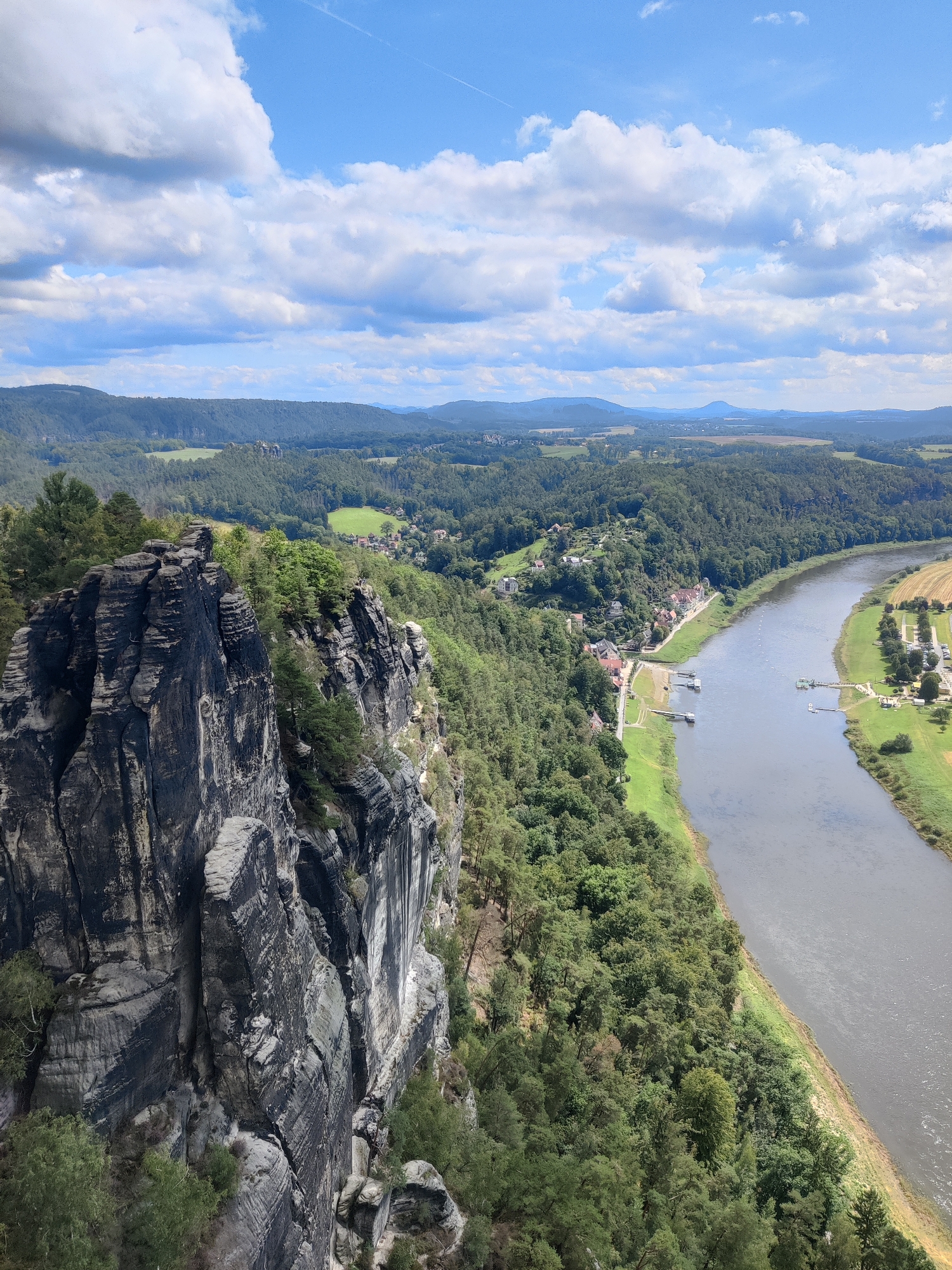This is a high-angle, landscape photograph taken from a rocky cliff, overlooking the Elbe River and the surrounding Saxon Switzerland National Park near the town of Lohmen, Germany. In the foreground, a prominent, dark grey, weathered rock formation juts out, its surface showing distinct layering and cracks. Green vegetation, including trees and bushes, grows on the slopes and along the edges of the cliff.
The middle ground is dominated by the wide, grey Elbe River, flowing from left to right across the frame. Along the riverbank, on the left side, a small village with red-tiled roofs and white buildings is nestled amongst the dense, green forest. There are visible docks and boats on the river in this area. On the right bank of the river, a strip of mown green grass leads to a few buildings and a parking area. The riverbanks are lined with green grass and trees, with agricultural fields visible further in the distance on the right.
The background consists of rolling, heavily forested hills that fade into the distance, with a hazy mountain range on the far horizon under a bright blue sky filled with scattered white clouds. A faint contrail from an aircraft is visible in the upper sky. The overall scene depicts a natural landscape with a river valley, forest, and a small settlement, viewed from a dramatic vantage point. There are no visible people in the image.