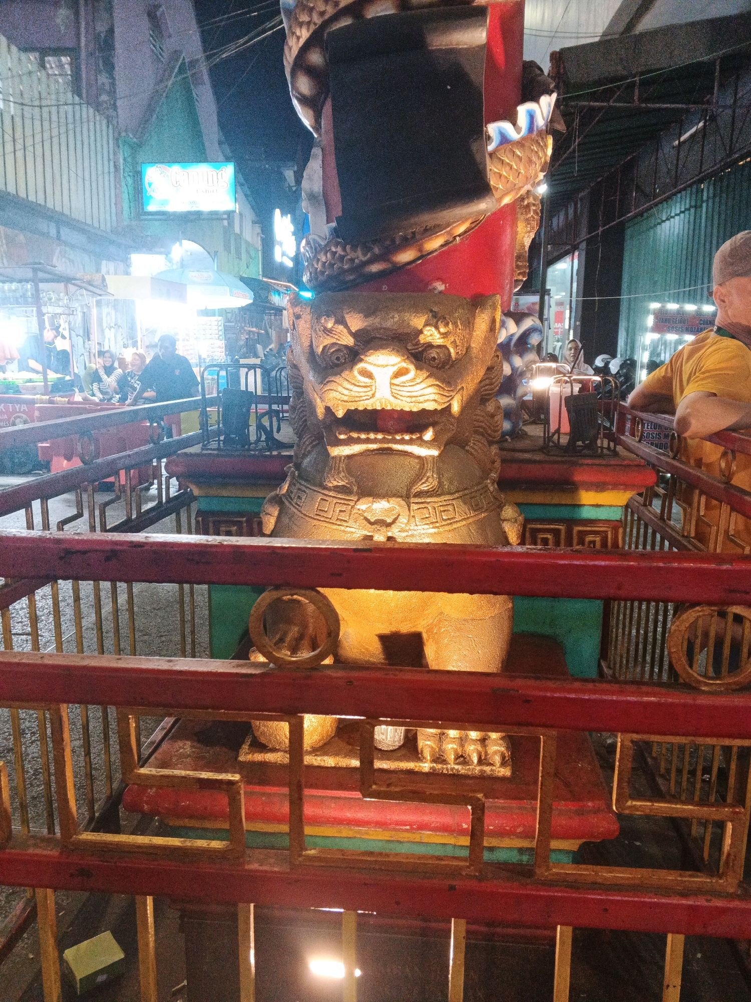 The image shows a close-up of a golden lion statue, likely a Foo dog, positioned behind a red and yellow railing. The statue has intricate details and is adorned with a large, dark headdress with red accents. The background suggests an outdoor marketplace or street scene at night, with neon signs and stalls visible. There are people in the background, some appearing to be shoppers or vendors. The overall impression is that of a festive or cultural event taking place in a bustling urban environment, possibly related to Chinese New Year or a similar celebration in Yogyakarta, Indonesia, given the presence of traditional elements like the lion statue.